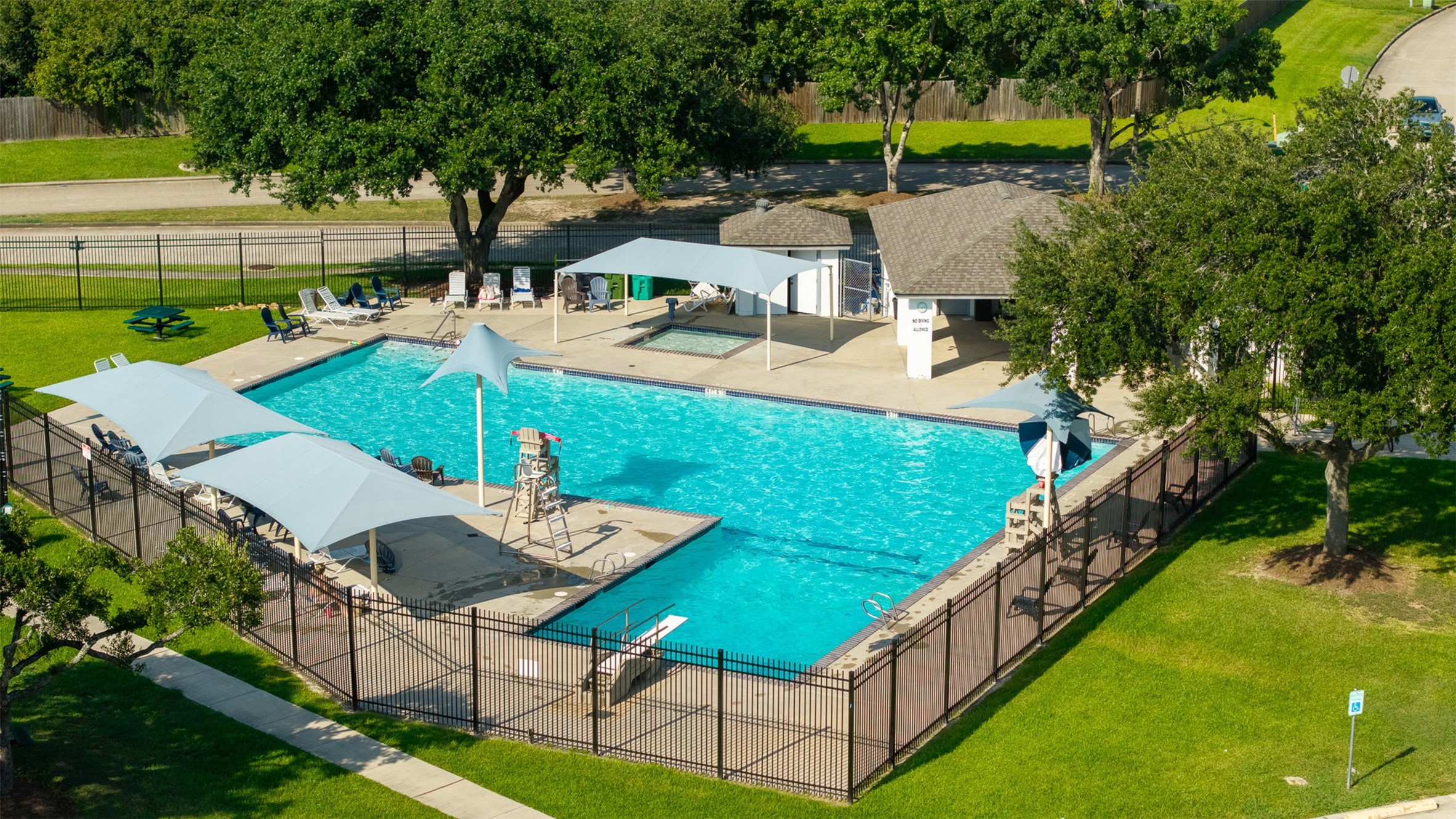 High-angle aerial capture showcases the expansive community pool area, complete with a shade canopy, lounge seating, and an adjacent splash pad. Residents enjoy this premier neighborhood amenity along with nearby sports courts and parks.