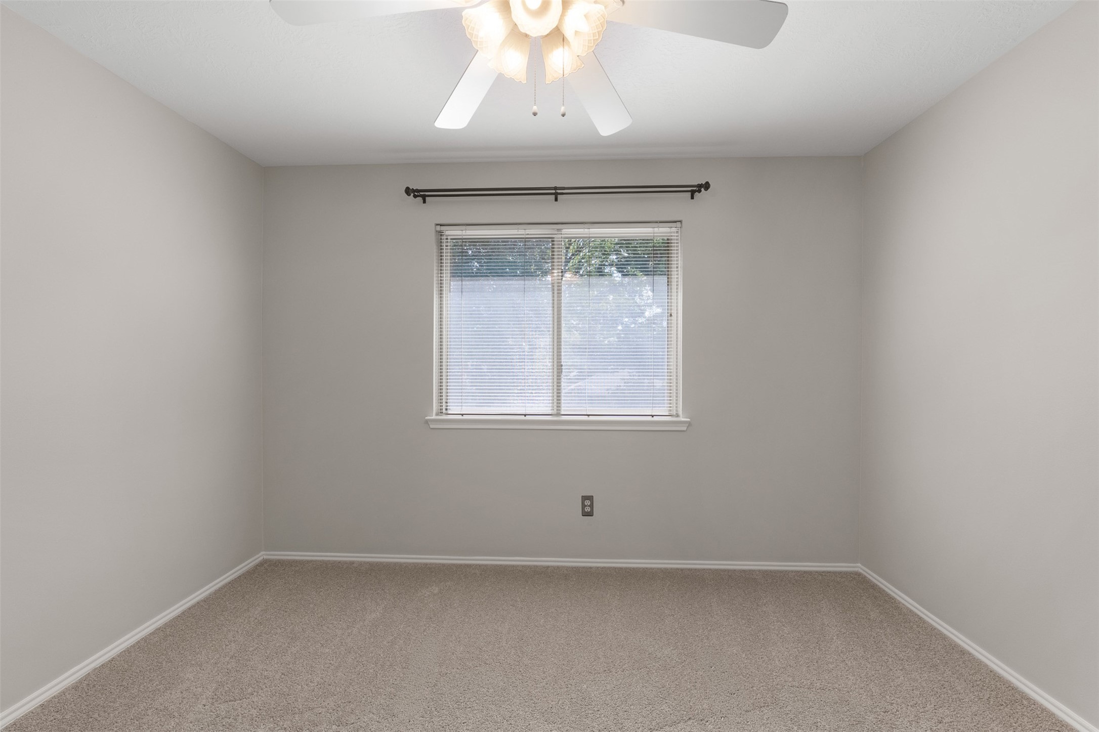 Soft carpet flooring and neutral walls define this secondary bedroom, anchored by a central window framed with blinds. A classic ceiling fan provides steady breeze and illumination in this versatile space.