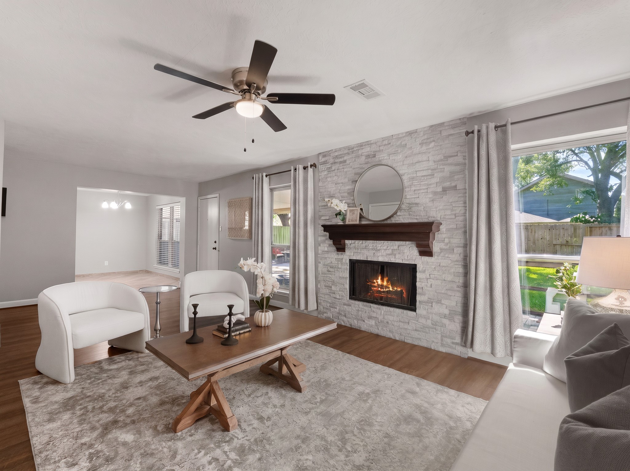 A focal stone stack gas fireplace framed by rich wood mantel accents anchors this open living area. Expansive windows frame green backyard views, creating a seamless connection to the nearby dining space.