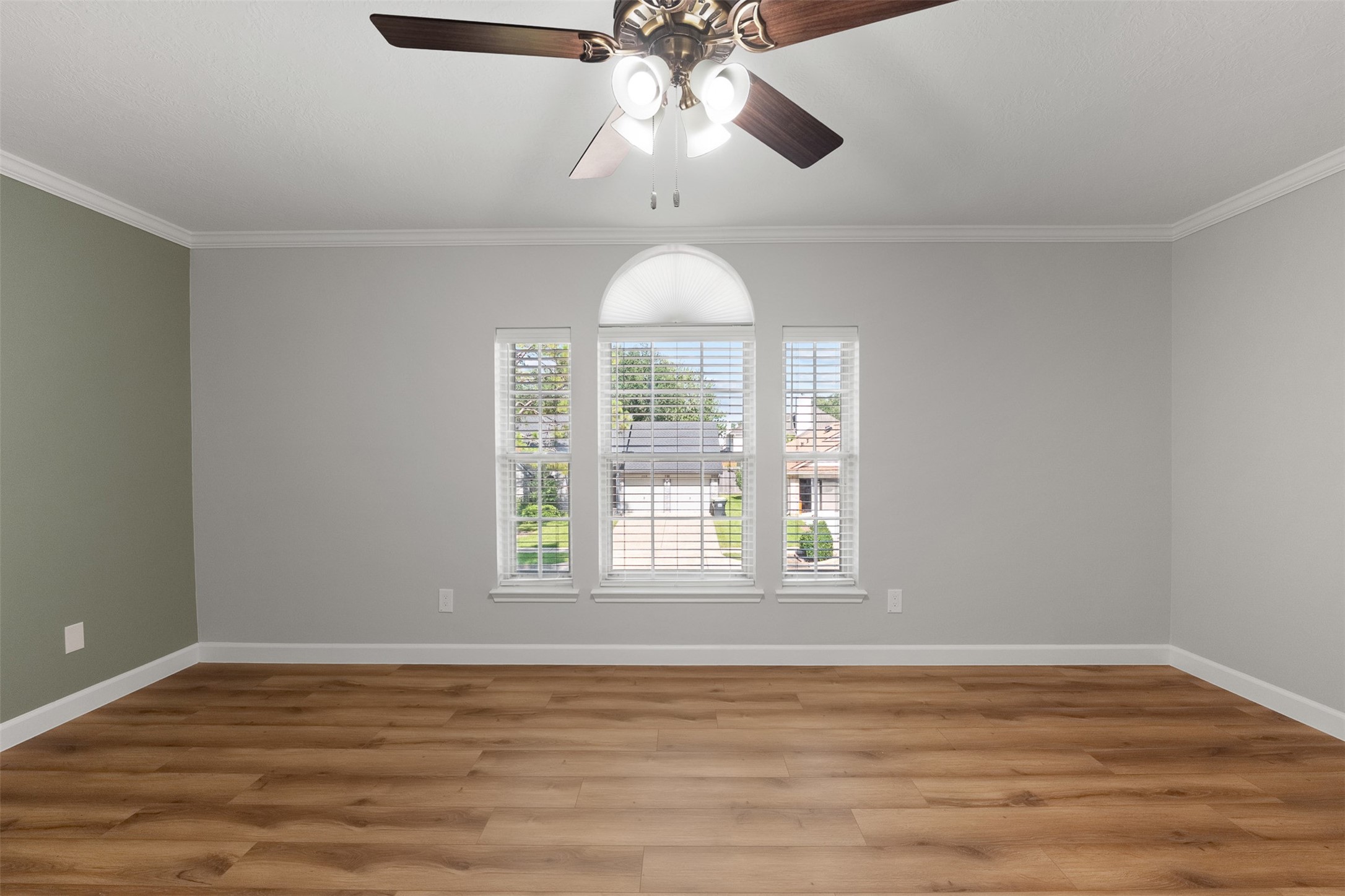 An arched center window framed by classic blinds serves as the focal point of this secondary bedroom, filling the room with light. Fresh neutral paint, crown molding, a sage green accent wall, and warm wood-look flooring combine to create a versatile space.