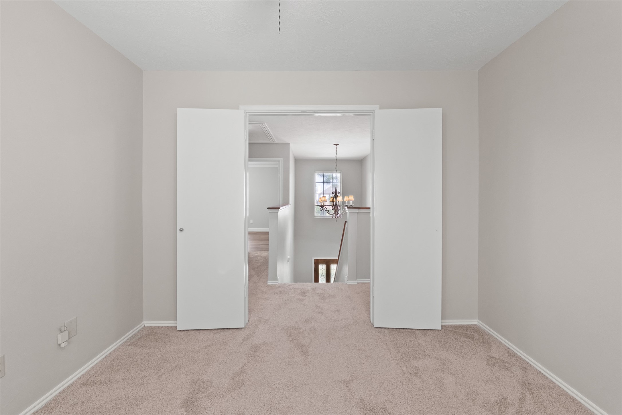 Double doors open to reveal this versatile secondary bedroom featuring fresh interior paint, soft carpet flooring, and a clear view out toward the upper landing chandelier.