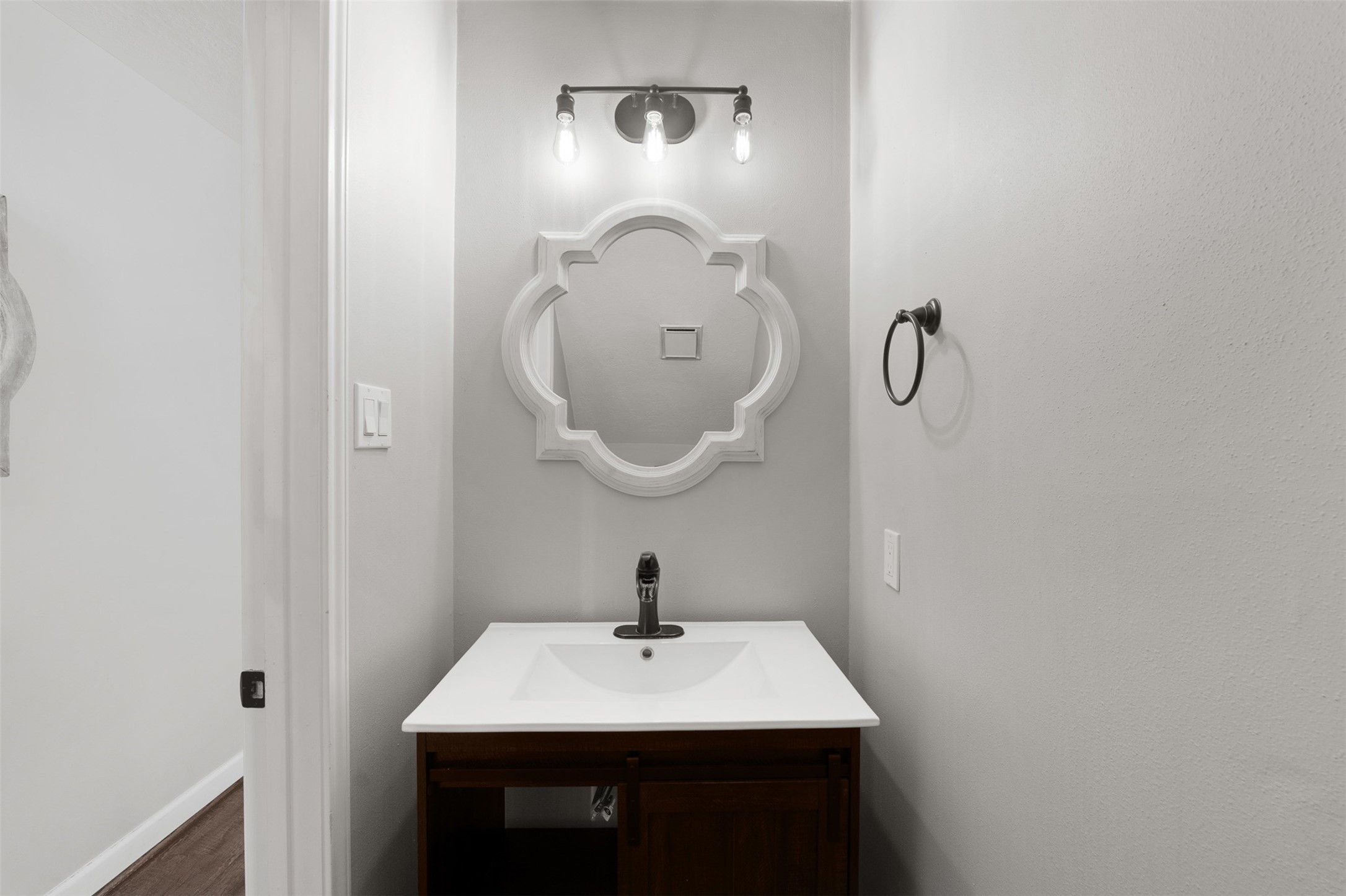 Featuring a renovated vanity with a white countertop and dark wood cabinetry, this half bath boasts a white quatrefoli mirror and vintage-inspired three-light sconce. Modern dark hardware and neutral walls create a polished, functional space for guests.