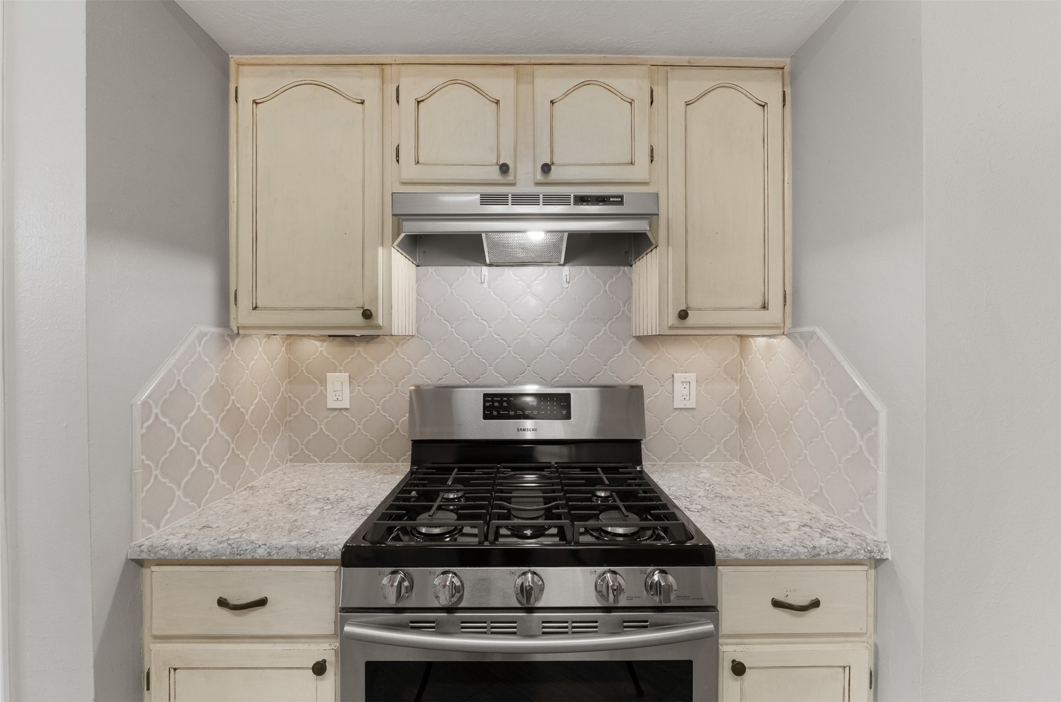 Featuring a gas range with an updated range hood, this kitchen highlight showcases an intricate arabesque designer backsplash flanked by distressed cream cabinetry. Under-cabinet lighting illuminates the quartz countertops, providing both functionality and style.