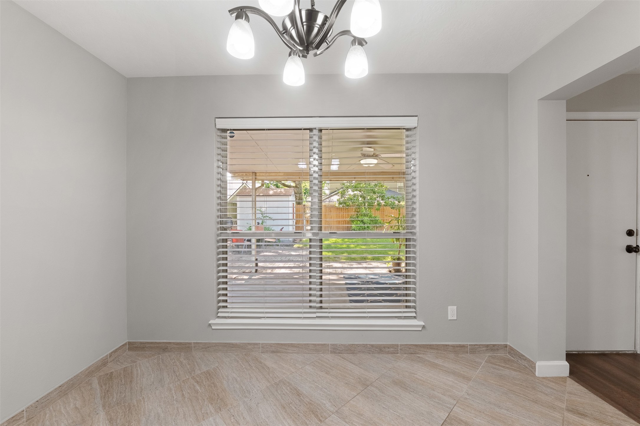 Positioned directly off the kitchen, this sunlit breakfast nook showcases diagonal tile flooring and a brushed nickel chandelier. A expansive window framed with blinds overlooks the covered patio and lush backyard, offering a serene backdrop for morning meals.