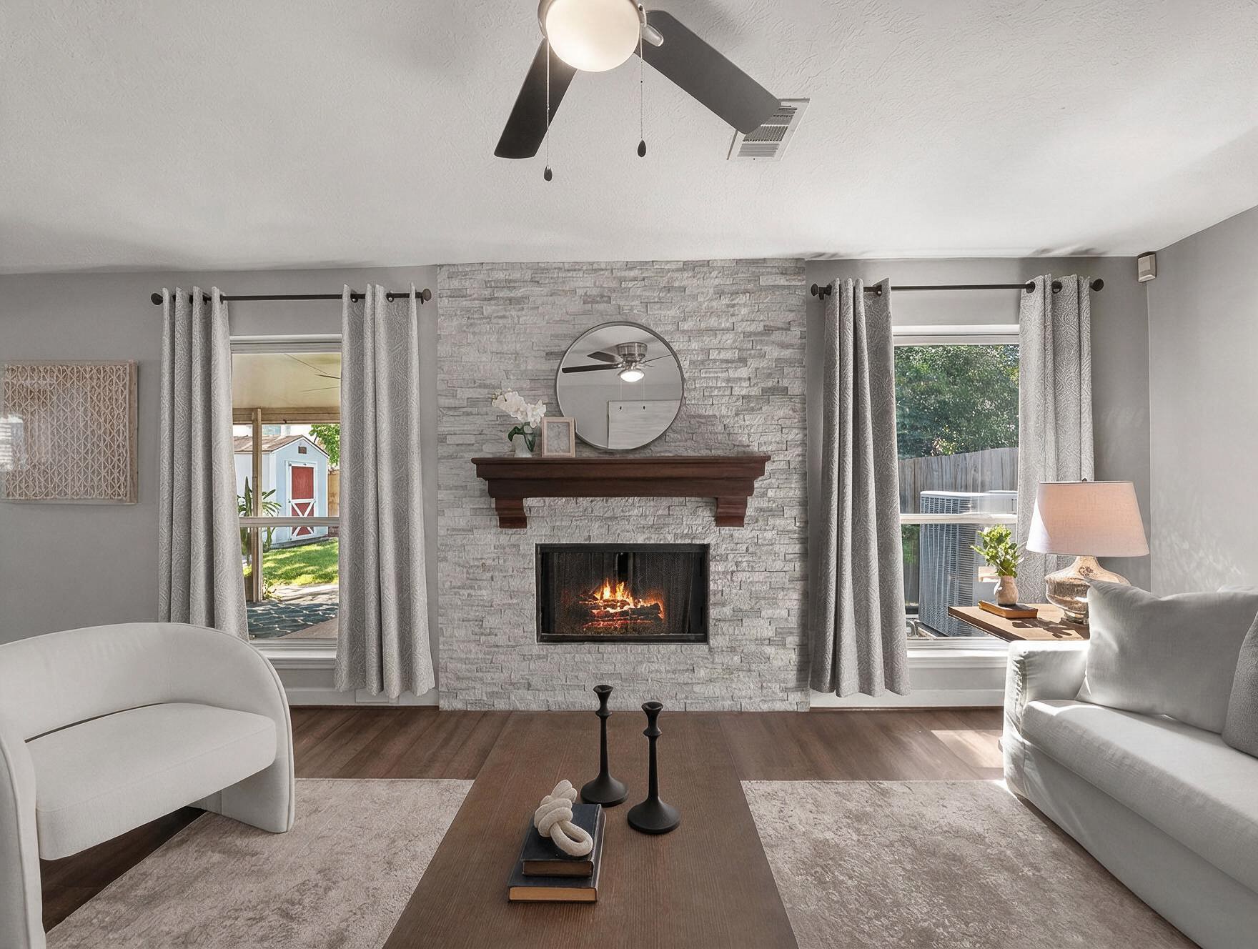 Flanked by tall windows, a magnificent stack stone fireplace serves as the centerpiece of this inviting living room. Rich wood-look flooring and plush neutral seating complement the stone hearth, while double windows offer direct views and access toward the covered outdoor patio.