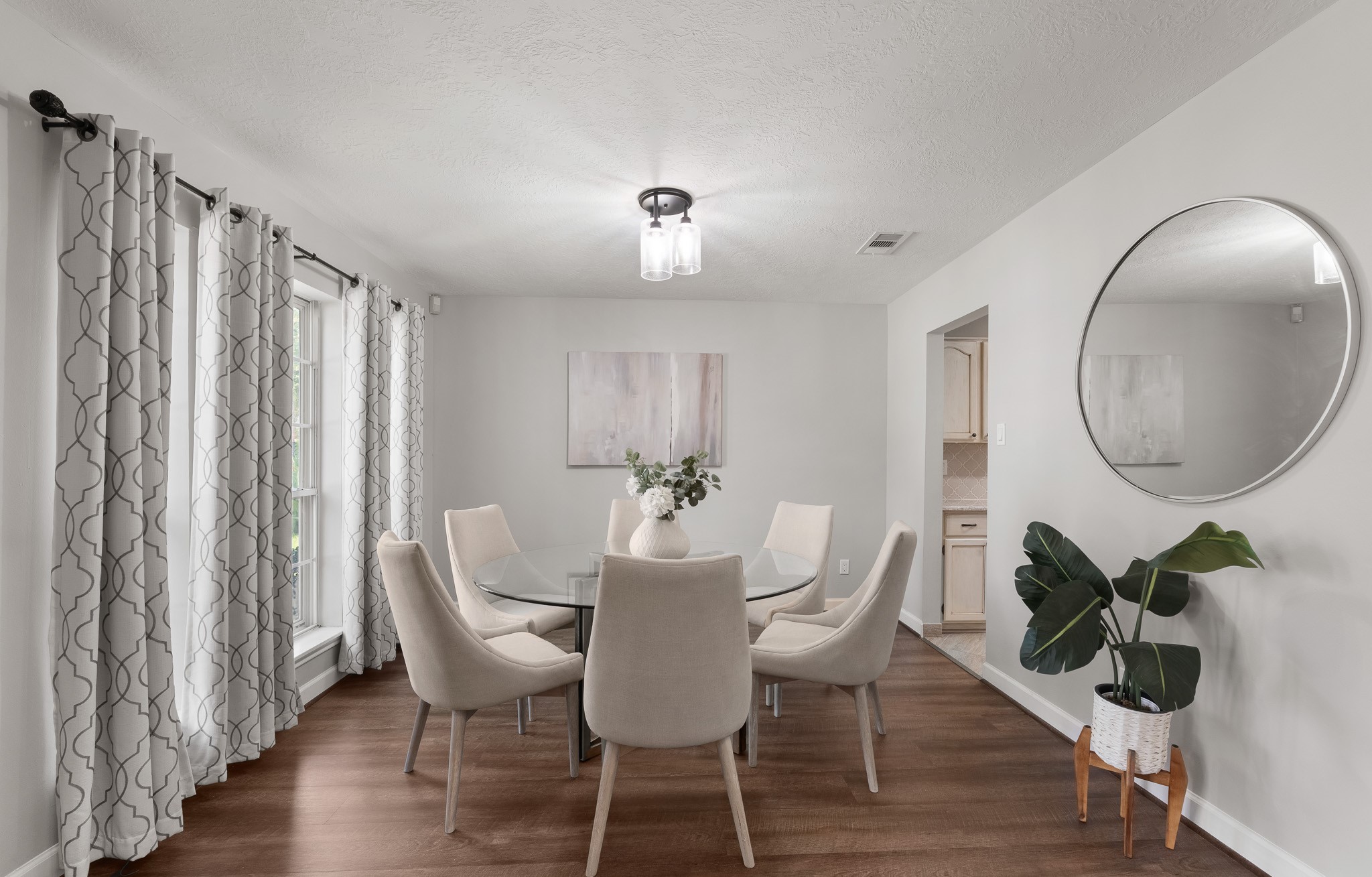 Sunlight streams through large windows into this refined formal dining room, highlighting rich wood-look flooring and a modern light fixture. The open layout accommodates a six-person table while offering seamless access to the adjacent kitchen.