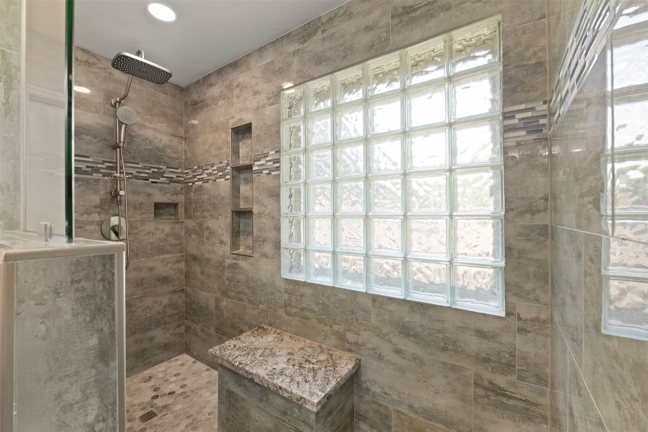 Framed in large-format grey tile, this renovated walk-in shower features a rain shower head, built-in bench with granite top, and an inset storage niche. Glass block window provides soft natural light while maintaining complete privacy.