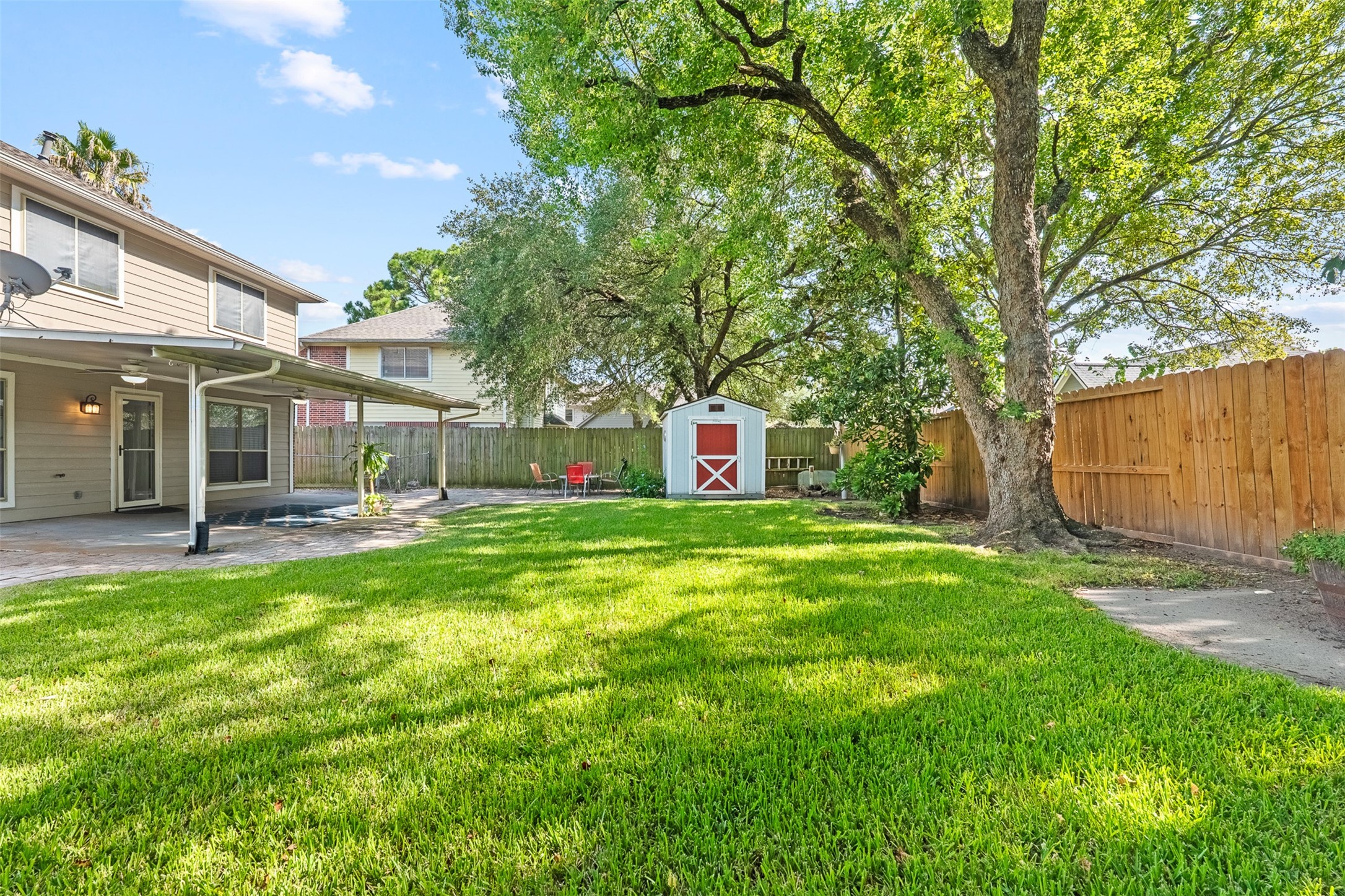 Expansive green lawn stretches past a classic red storage shed, towering shade trees, and a full privacy fence in this tranquil backyard. A paver extension off the covered patio provides easy access for outdoor entertaining and relaxation.