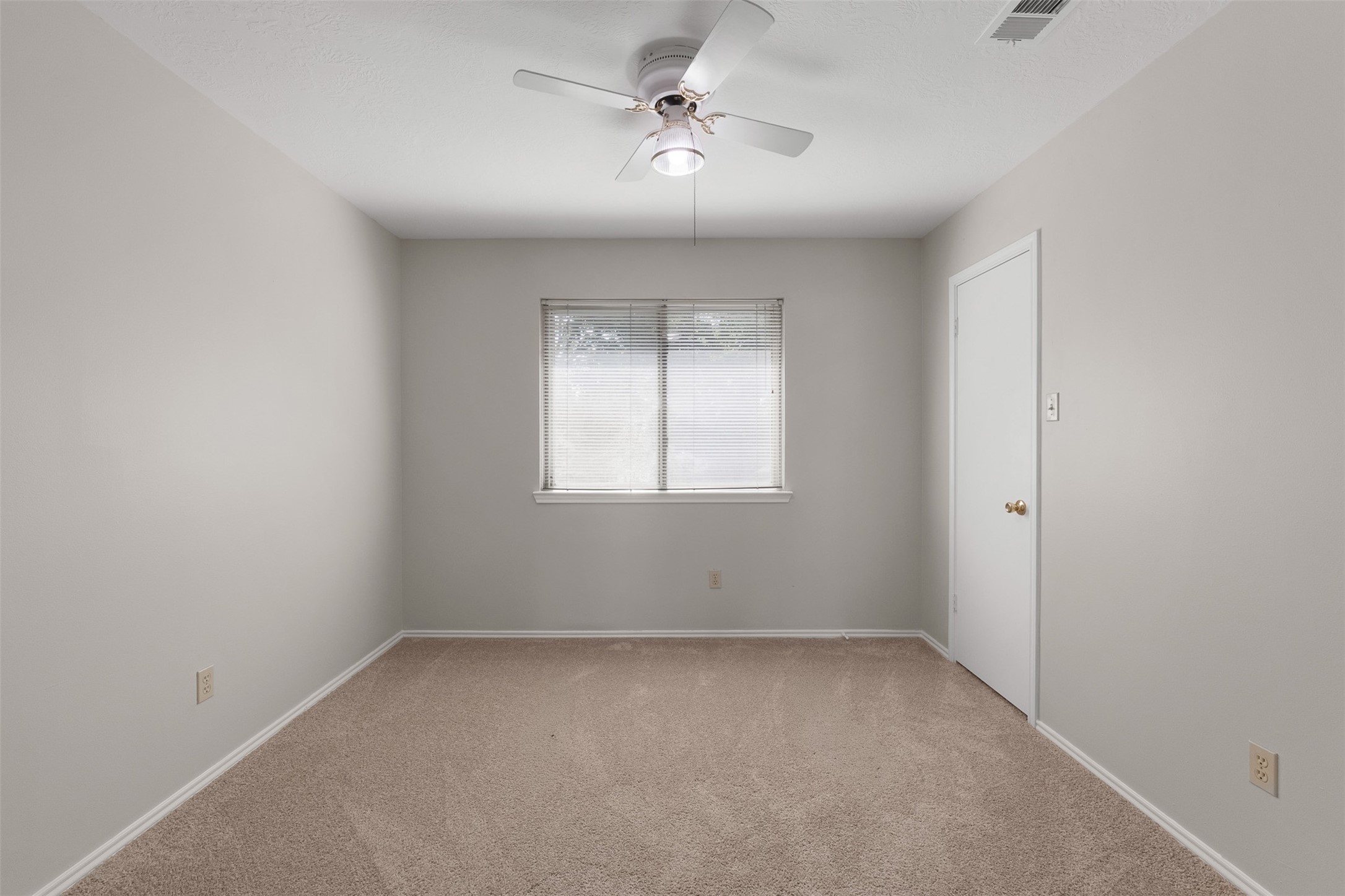 Neutral walls and cozy carpet flooring define this secondary bedroom, anchored by a central window with blinds. A traditional ceiling fan provides comfortable illumination, while fresh interior paint creates a bright atmosphere.