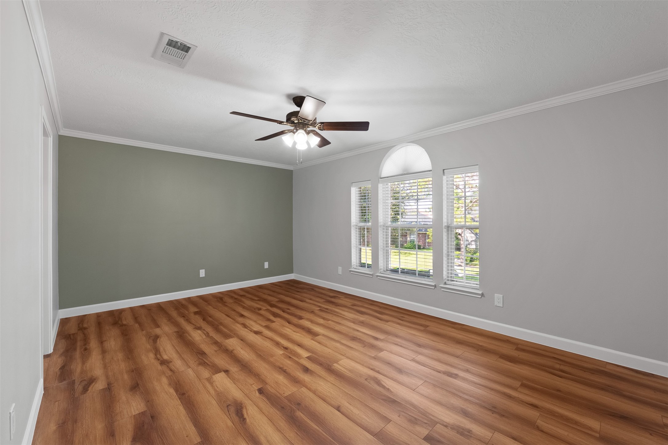 Warm wood-look flooring stretches across this light-filled room, framed by crisp crown molding and a sage green accent wall. Wide windows with a center arch fill the space with light, while a dark wood ceiling fan provides steady breeze and brightness.