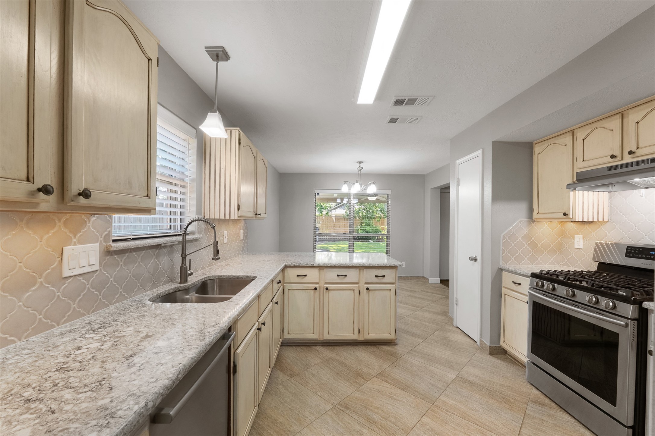 Featuring quartz countertops and a designer backsplash, this kitchen showcases a gas cooktop, dual sink, and cream cabinetry with ample storage. The functional peninsula extends into a breakfast bar, seamlessly connecting to the sunlit breakfast area framed by oversized windows.