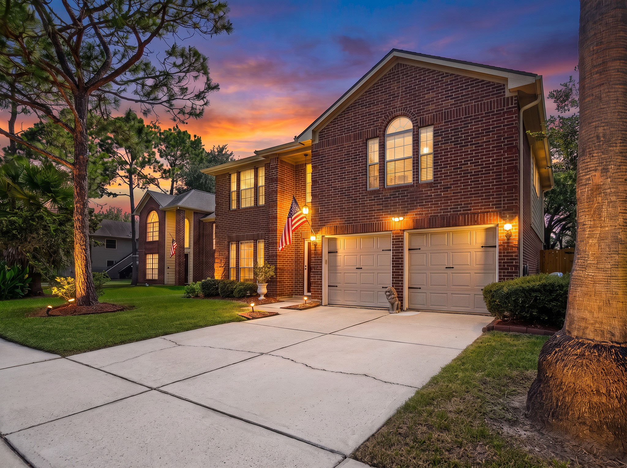 A wide concrete driveway leads to the two-car attached garage of this classic two-story brick home, framed by lush front lawn landscaping and accent illumination.