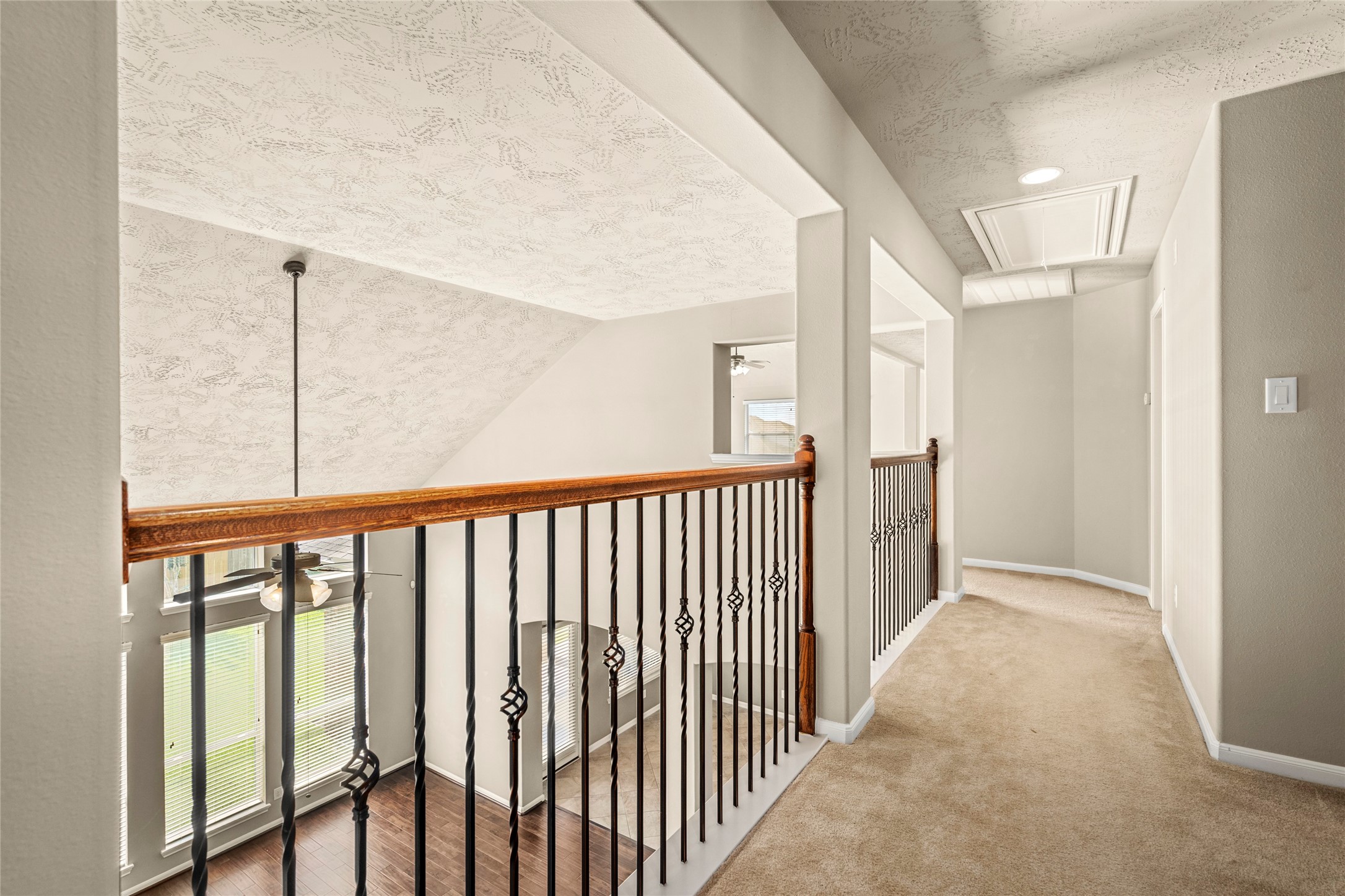 This showcases the bright, open hallway with a modern metal and wood railing overlooking a two-story living area. The space features neutral carpeting and textured ceilings, creating a warm and inviting atmosphere. GOING TO THE GAME ROOM AND ROOMS