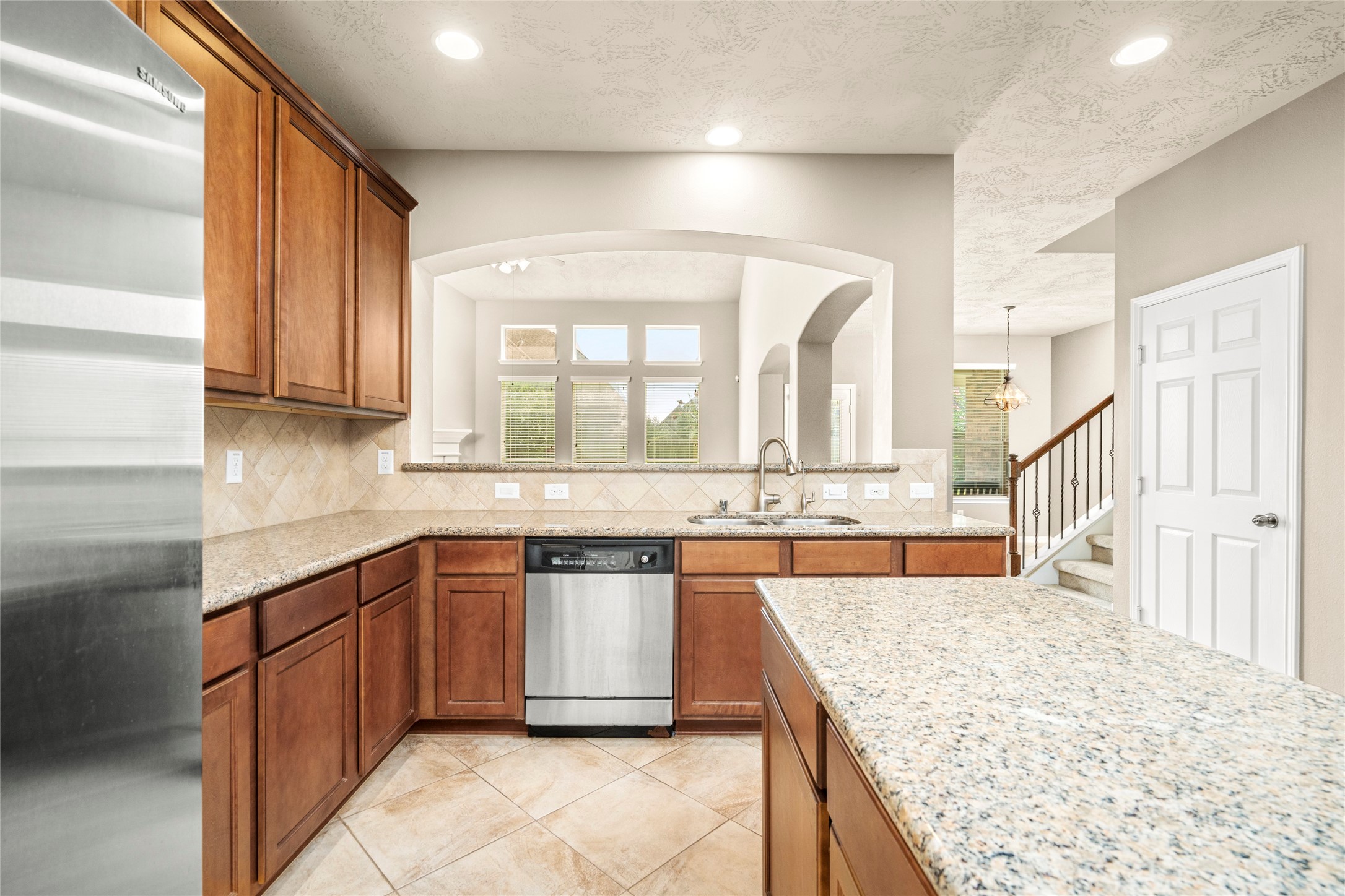This kitchen features granite countertops, wooden cabinetry, and stainless steel appliances. It opens to a bright living area with large windows and an arched pass-through, enhancing the spacious feel.