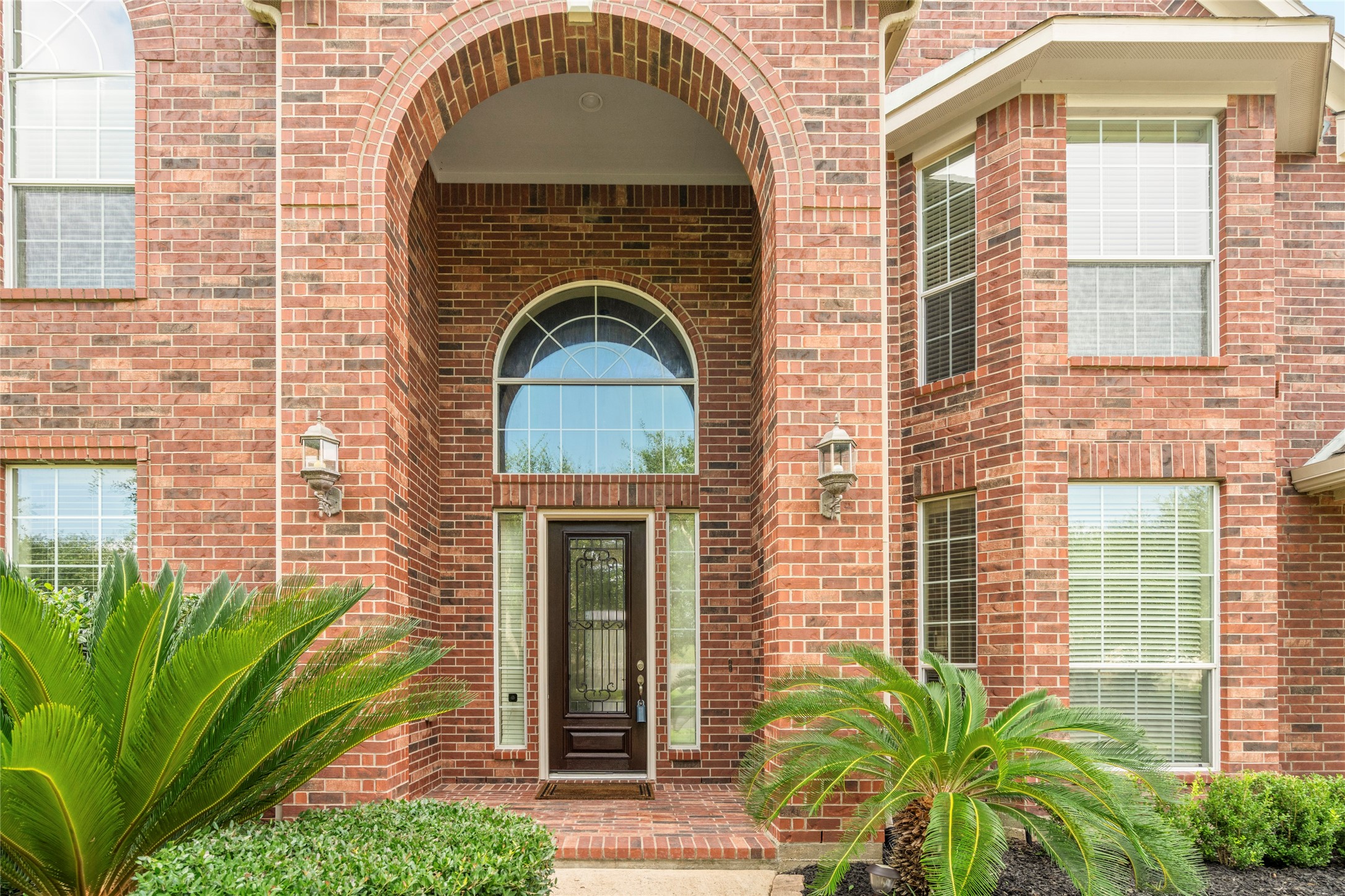 A breathtaking arched brick entryway sets a grand tone from the curb, framing a stately wood and wrought-iron front door beneath a sweeping arched transom window  striking craftsmanship and elegant landscaping welcome you home.