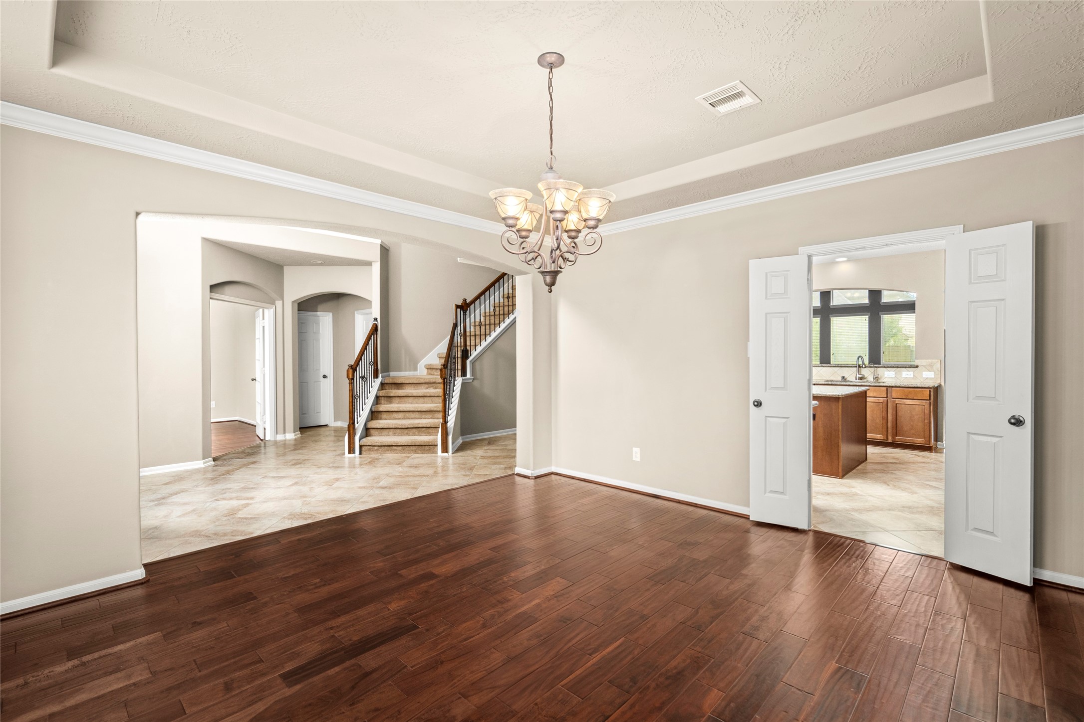 A seamless transition of rich wood and tile flooring leads from the dining room toward the staircase and kitchen, illuminated by a striking chandelier. Luxury living, beautifully connected.