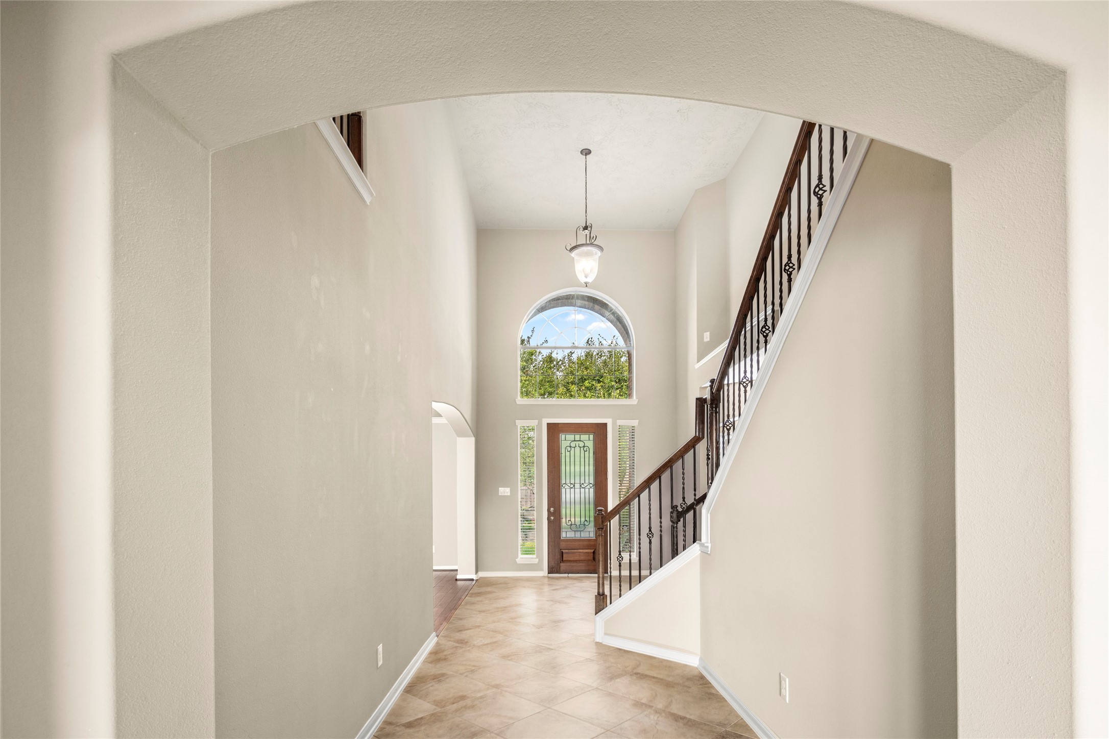 Step into pure elegance: a dramatic two-story foyer crowned by a sweeping arched window, anchored by a graceful staircase finished in rich dark wood and wrought iron, set atop sun-kissed tile flooring.