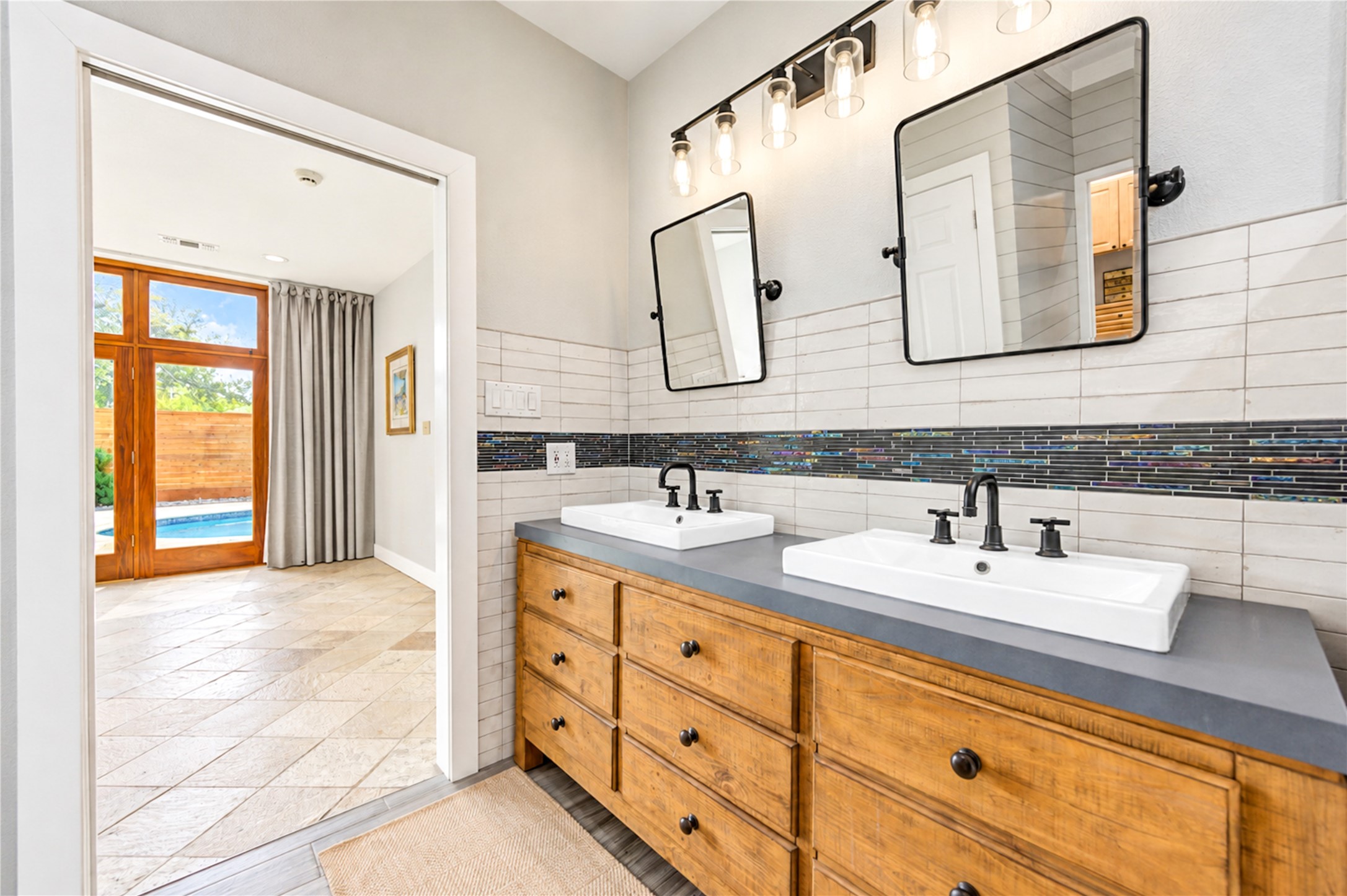 Linear white tile, a mosaic accent band and warm wood cabinetry frame the primary bath, with a view through to the glass doors of the primary bedroom.