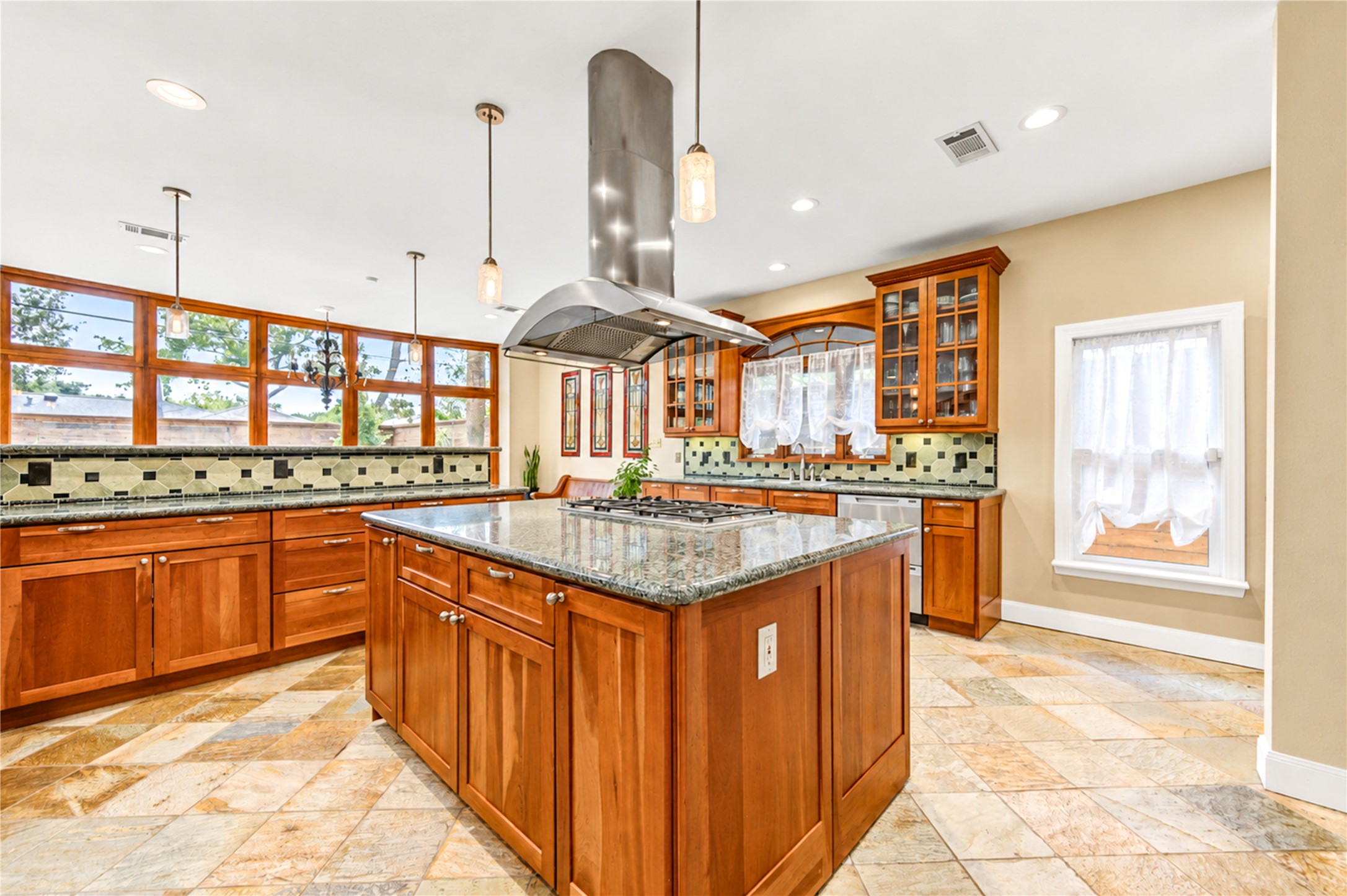 A gas cooktop island beneath the stainless vent hood anchors the kitchen, framed by a window wall and a decorative tile backsplash.