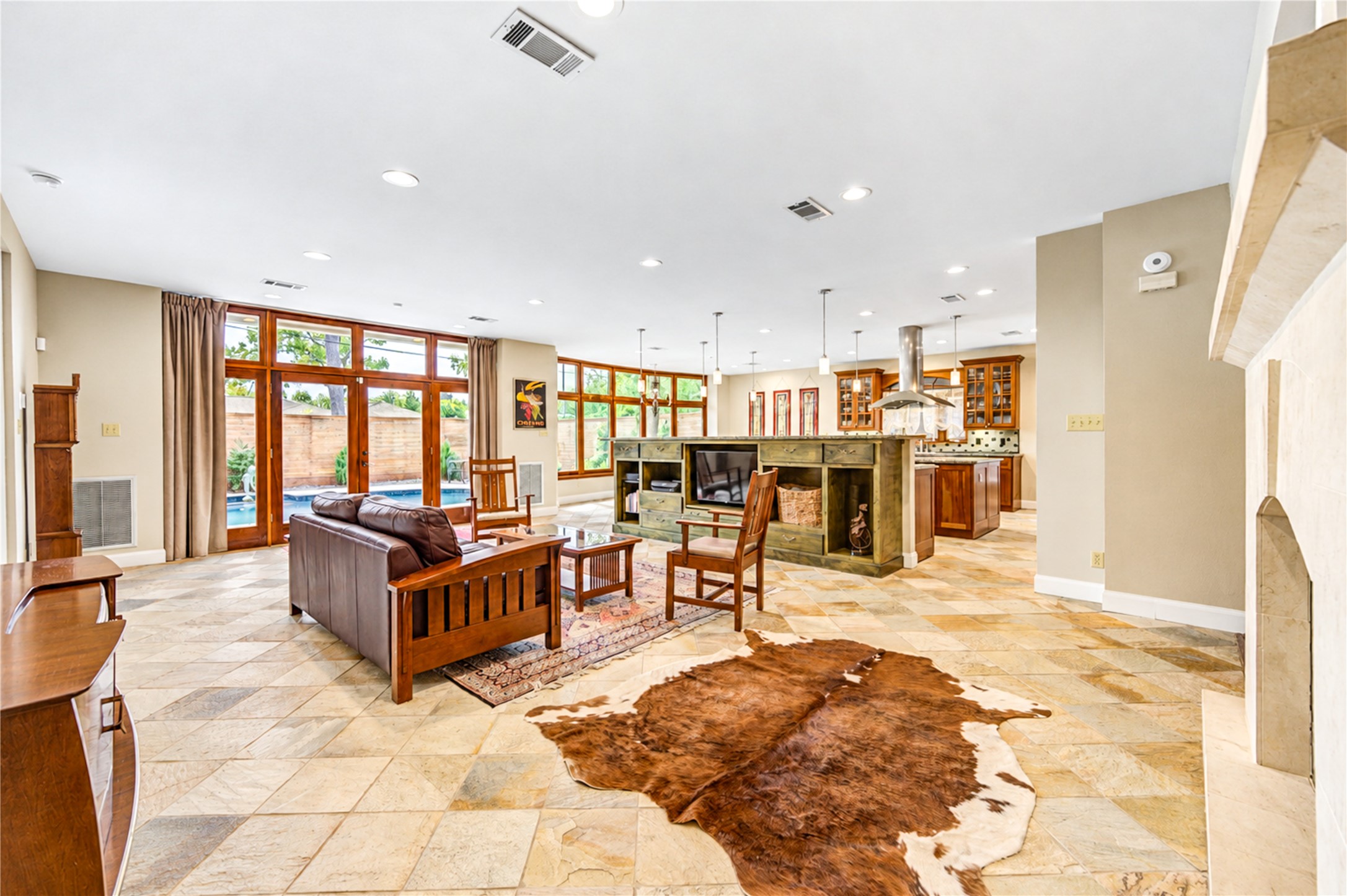 Stone flooring flows through the open family room and kitchen, with the fireplace on one side and wood-framed glass doors to the pool on the other.