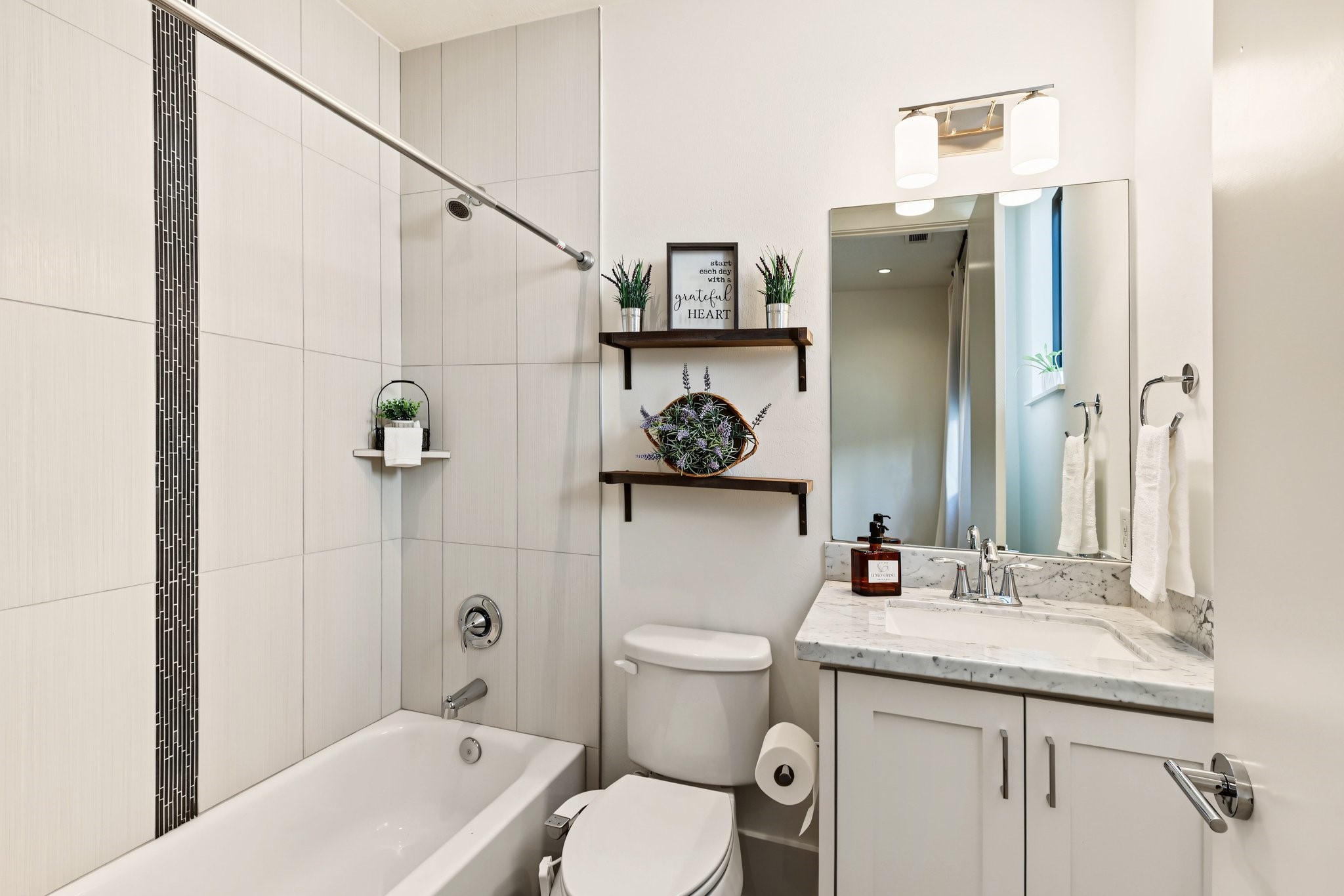 Unwind in a private en-suite bath where a deep soaking tub and full-height tile surround create a tranquil, spa-like setting. A contrasting dark mosaic accent adds tailored definition, while polished chrome fixtures complete the sleek, contemporary aesthetic.