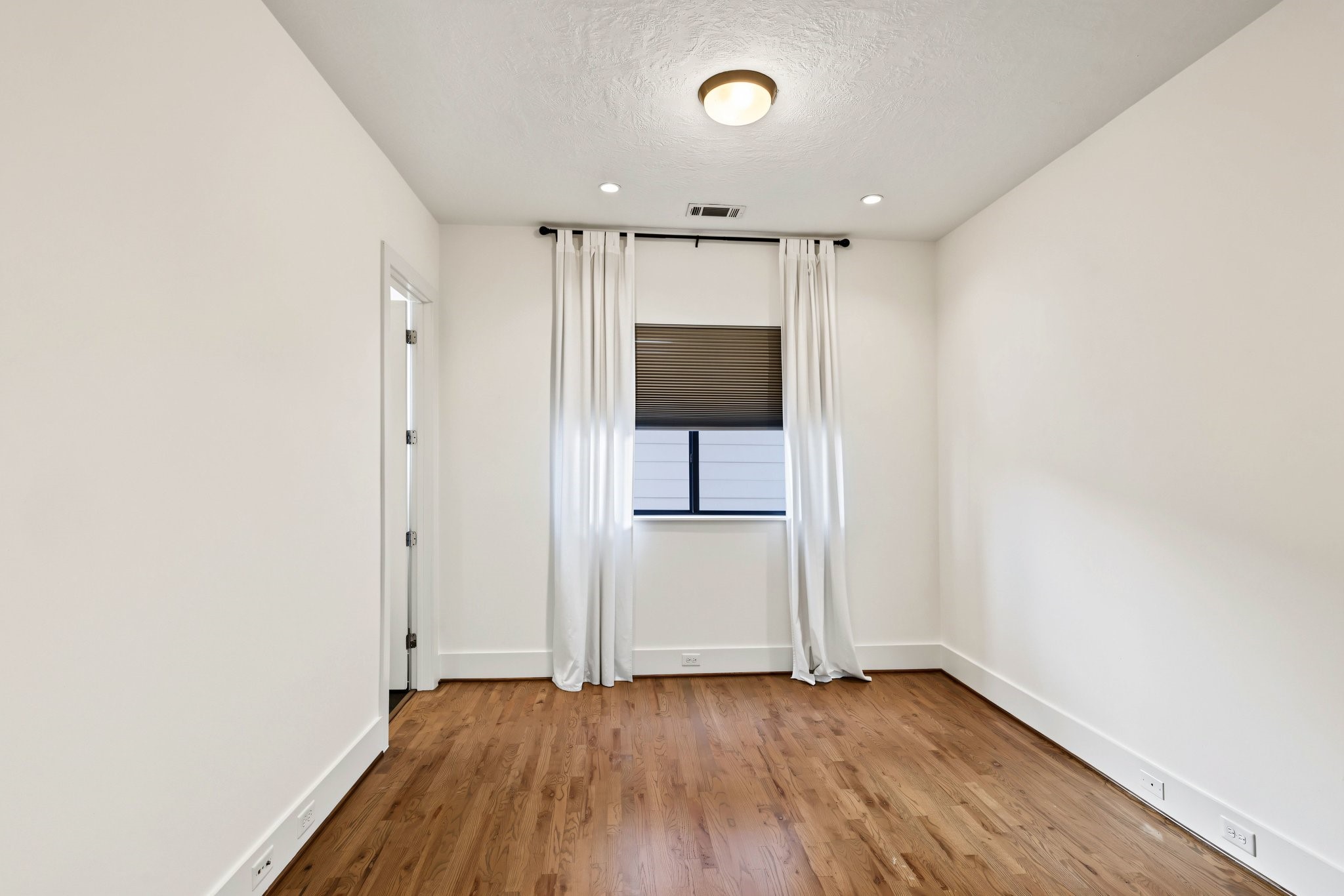 This secondary bedroom offers a sunlit, versatile retreat finished with warm natural hardwoods and crisp gallery-white walls. A central picture window features an accordion privacy shade paired with flowing, full-length white drapery on a contrasting dark rod, providing effortless light control and timeless style.