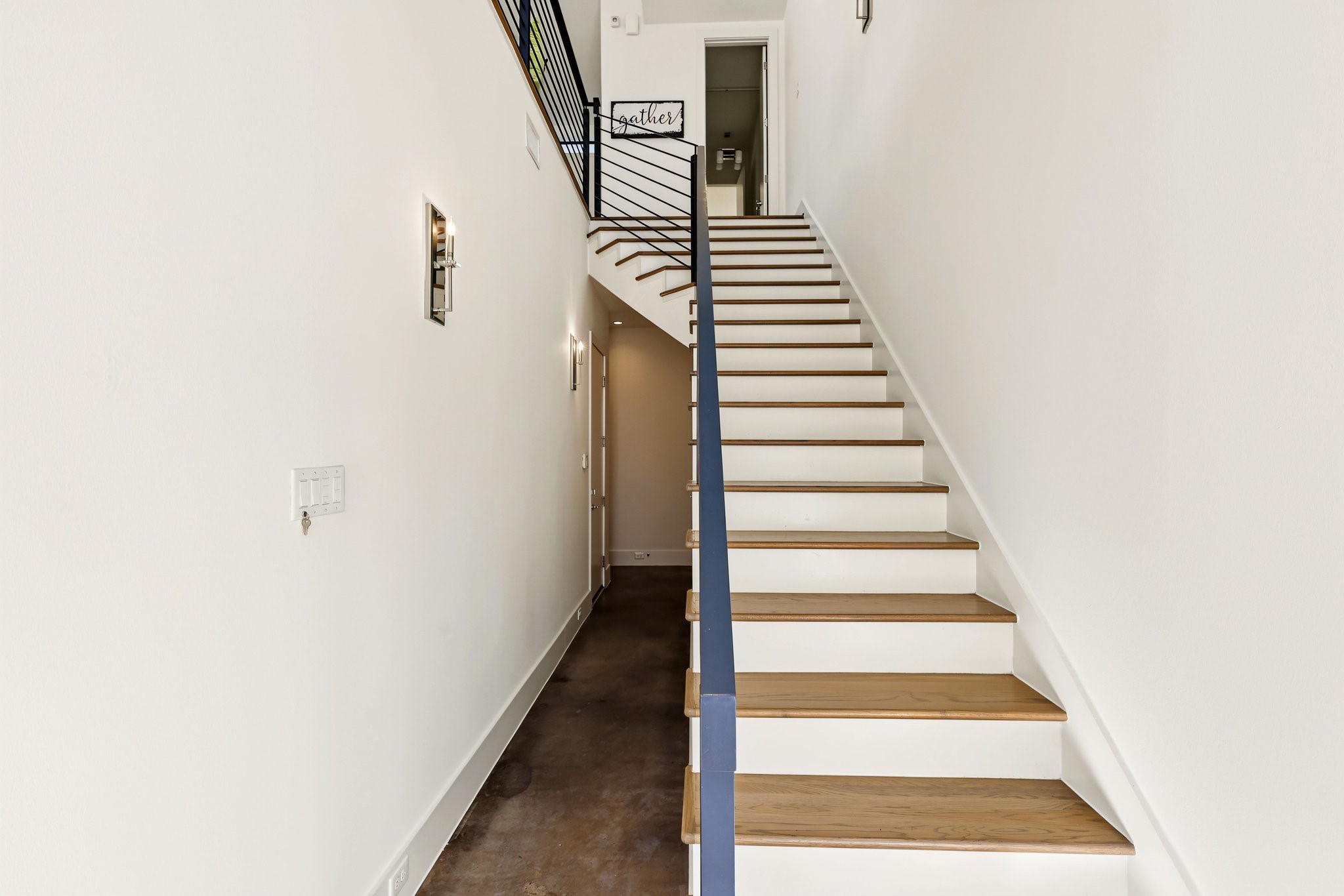 The interior entryway features a straight architectural staircase serving as the focal point, crafted with warm natural-wood treads contrasted against clean white risers. It is framed by a sleek, dark-finished banister and an open horizontal metal railing along the upper landing.