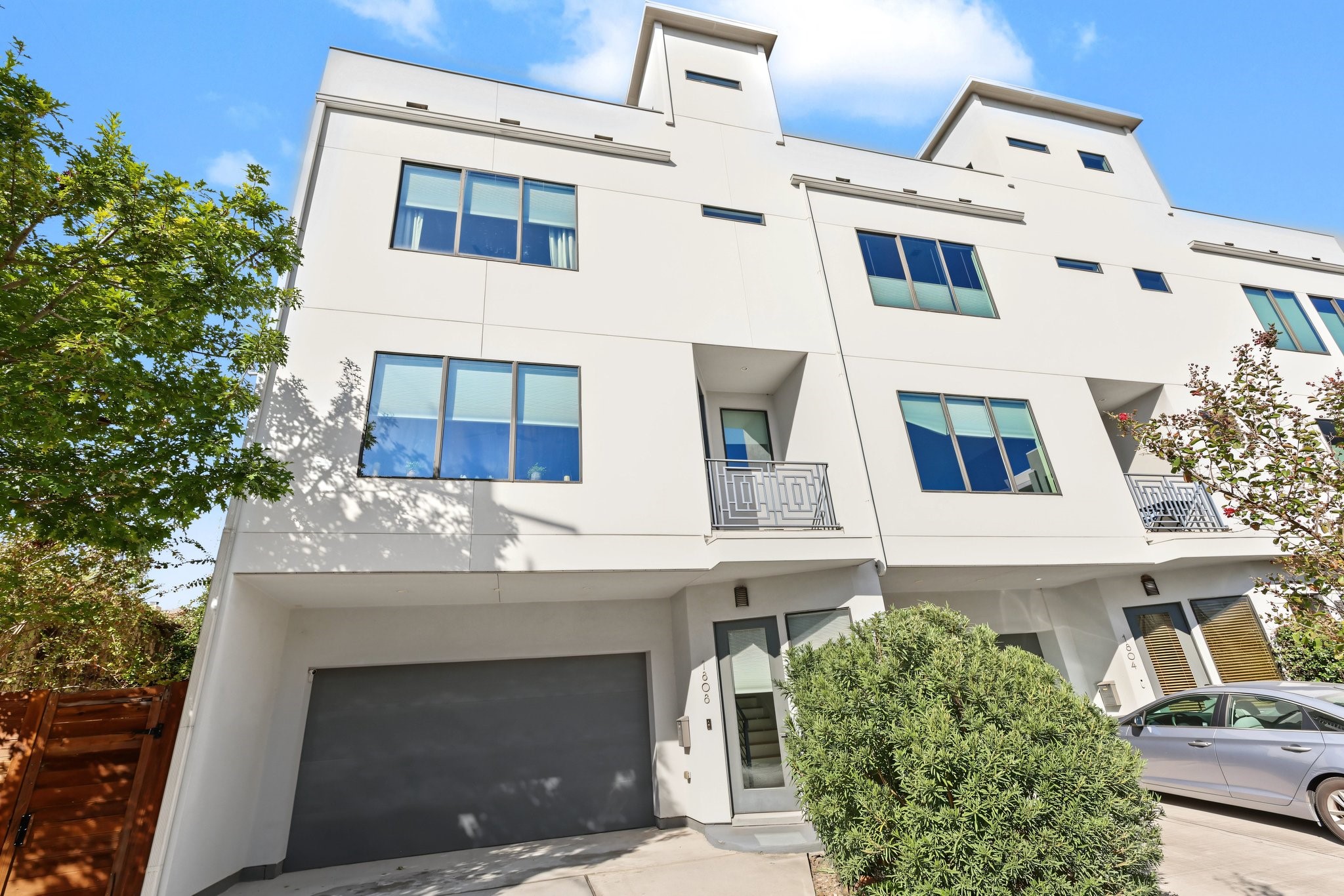 At ground level, a broad concrete driveway leads to a private, integrated garage finished with a sleek charcoal-toned overhead door. Adjacent to the garage, the sheltered entryway is flanked by an exterior sconce light and concrete walkway. The upper living levels elevate the facade with a recessed second-story balcony accented by a decorative geometric wrought-iron railing, offering an inviting outdoor vantage point.