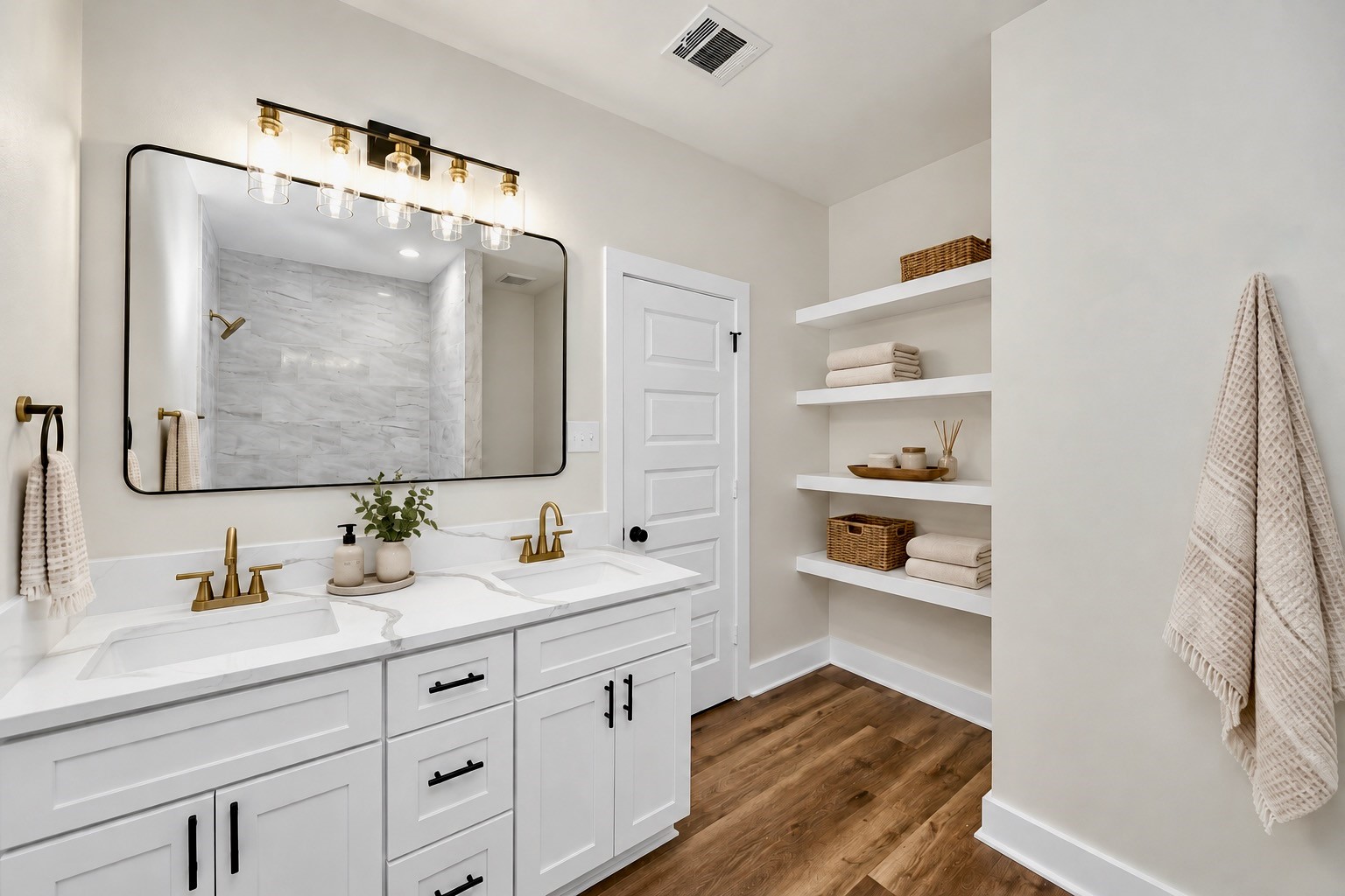 Primary bathroom featuring a double-sink vanity, quartz countertops, white cabinetry with modern hardware, stylish framed mirror, gold-tone fixtures & built-in shelving for additional storage.