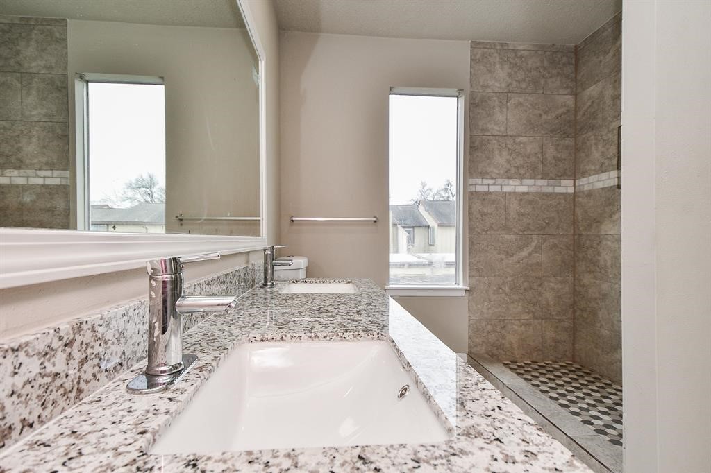 Modern bathroom featuring a large granite countertop with dual sinks, sleek fixtures, and a spacious walk-in shower with elegant tilework. Natural light fills the space through two tall windows.