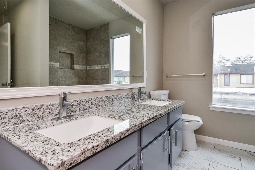 This bathroom features a sleek double-sink vanity with granite countertops, modern fixtures, and a large mirror. The room is brightened by a big window and includes a tiled shower area and a toilet.