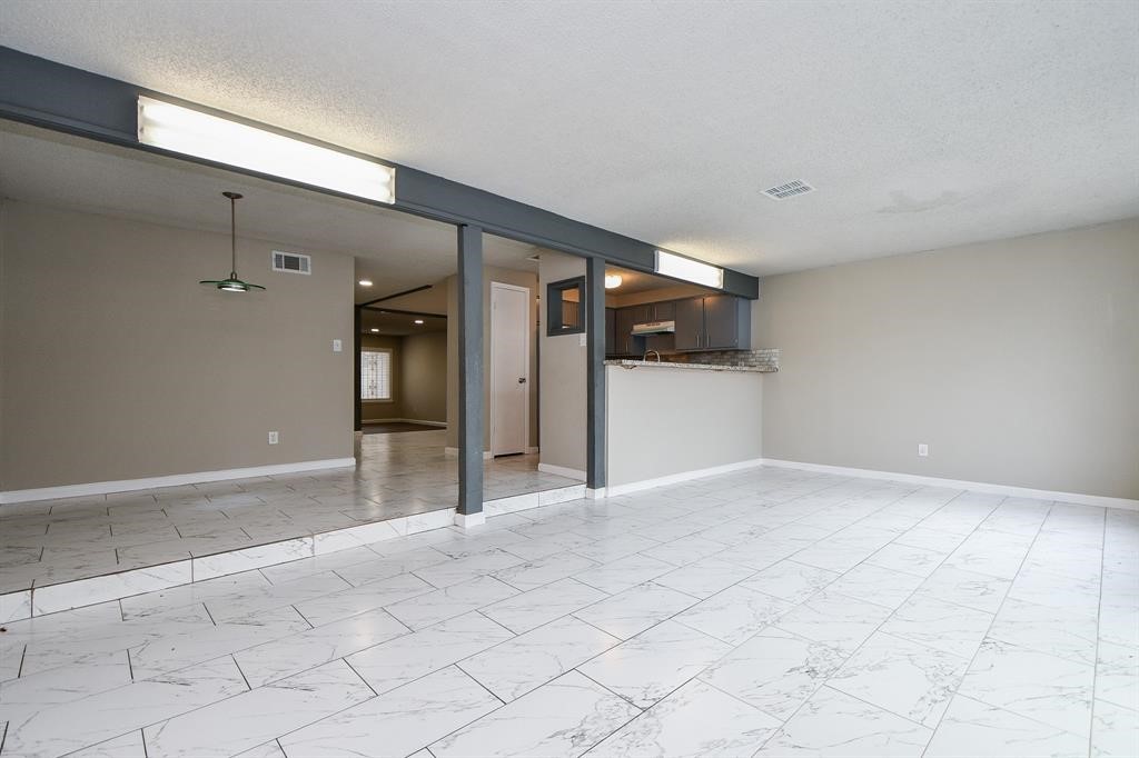 Spacious room with light-colored tile flooring and neutral walls, featuring an open layout that connects to a kitchen with a breakfast bar. Recessed lighting and a large window provide ample natural light.