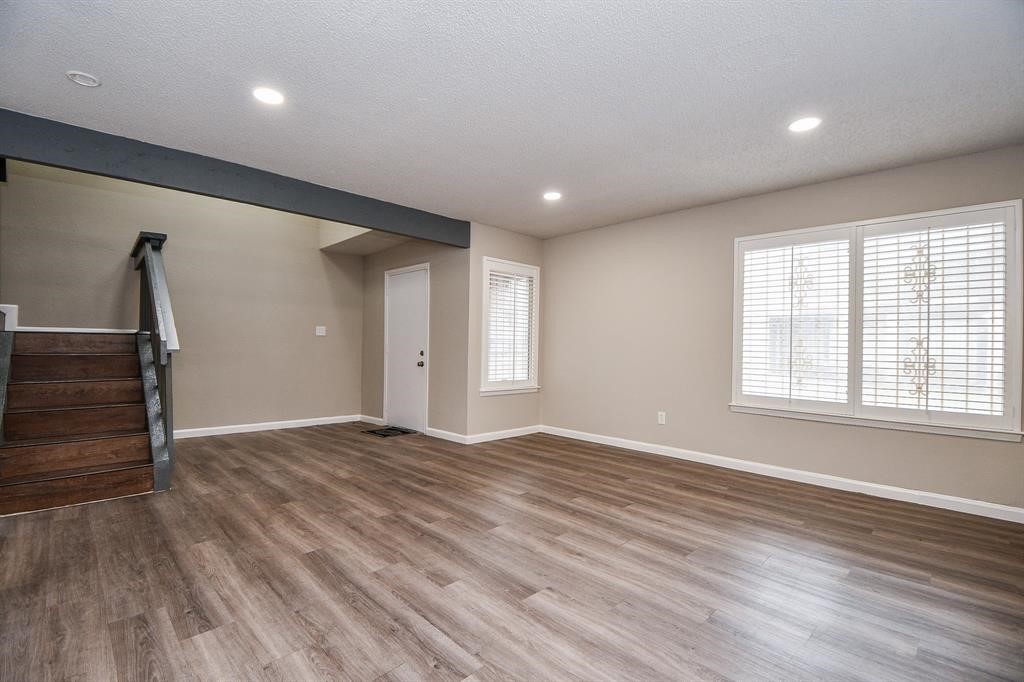 This spacious living area features modern laminate flooring, neutral walls, recessed lighting, and large windows with blinds. The dark wood staircase adds character, leading to the upper level, and there's a convenient side door for easy access.