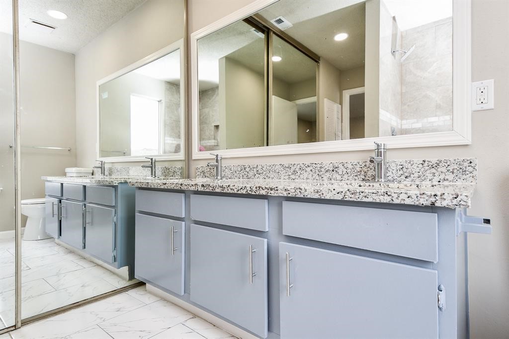 This bathroom features a dual-sink vanity with granite countertops, large mirrors, and light blue cabinetry. The space includes modern fixtures and a neutral color palette, complemented by a tiled floor and shower area.