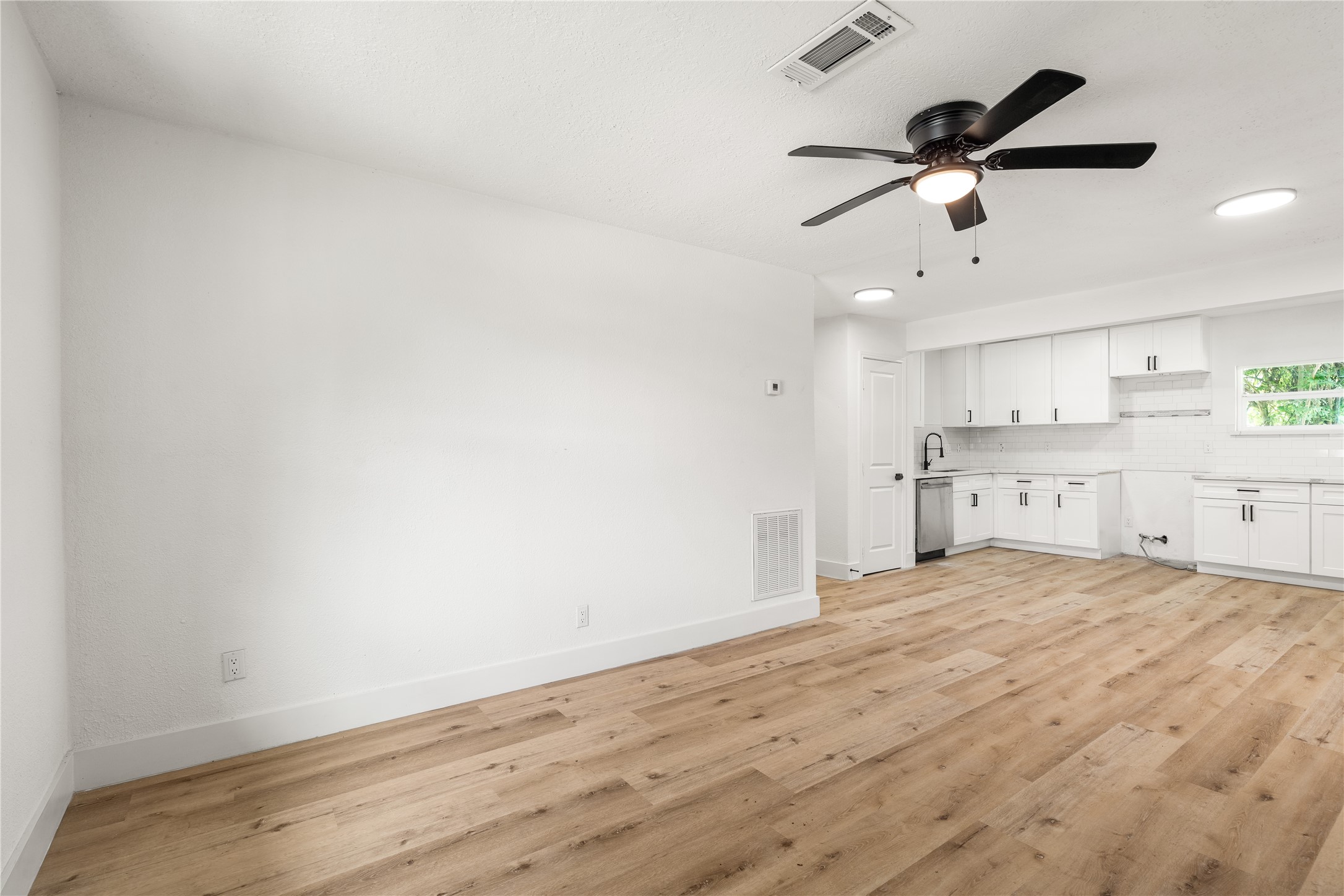 Wide-angle view taken from the main living room wall, showing side window light, warm wood-grain floors, and an open layout looking directly into the kitchen. Wide-angle view taken from the main living room wall, showing side window light, warm wood-grain floors, and an open layout looking directly into the kitchen.