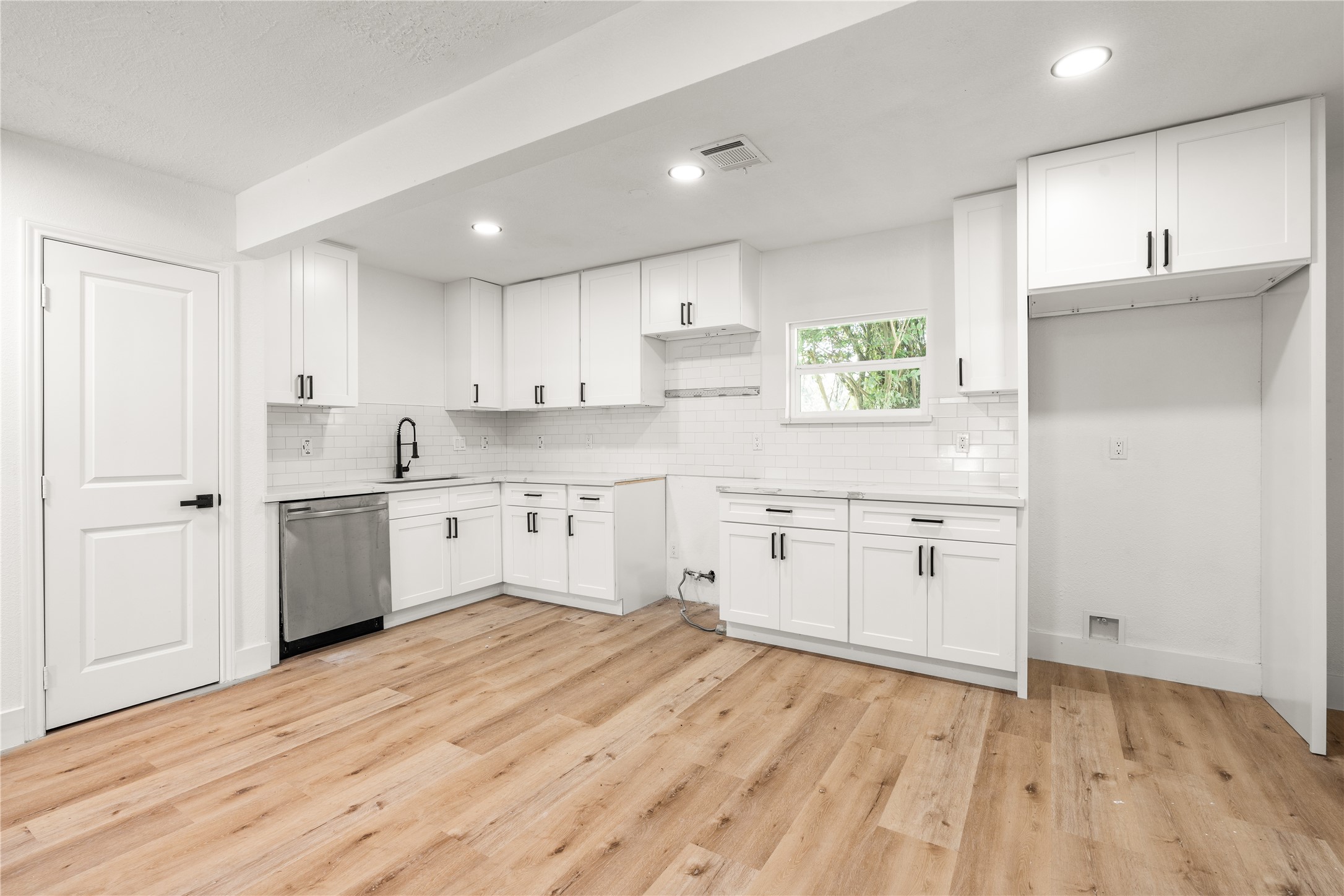 Full wide-angle view of the unstaged kitchen and dining area facing the main workspace, highlighting white shaker cabinets, matte black accents, subway tile, a stainless dishwasher, and continuous light wood-grain flooring. Full wide-angle view of the unstaged kitchen and dining area facing the main workspace, highlighting white shaker cabinets, matte black accents, subway tile, a stainless dishwasher, and continuous light wood-grain flooring.