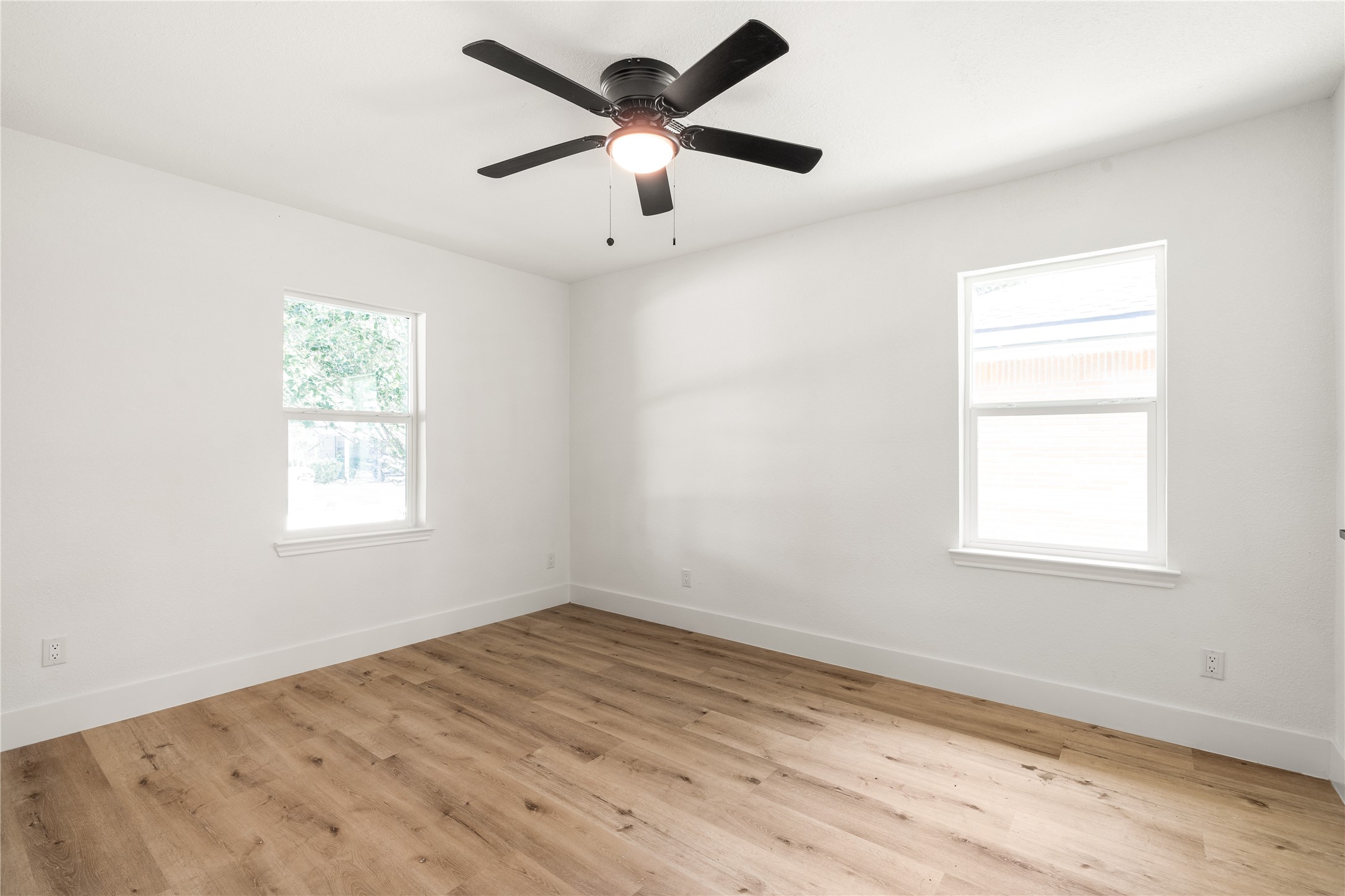 Bright bedroom corner featuring dual windows, modern ceiling fan, and warm wood-grain flooring. Ideal for a guest room or quiet home office.