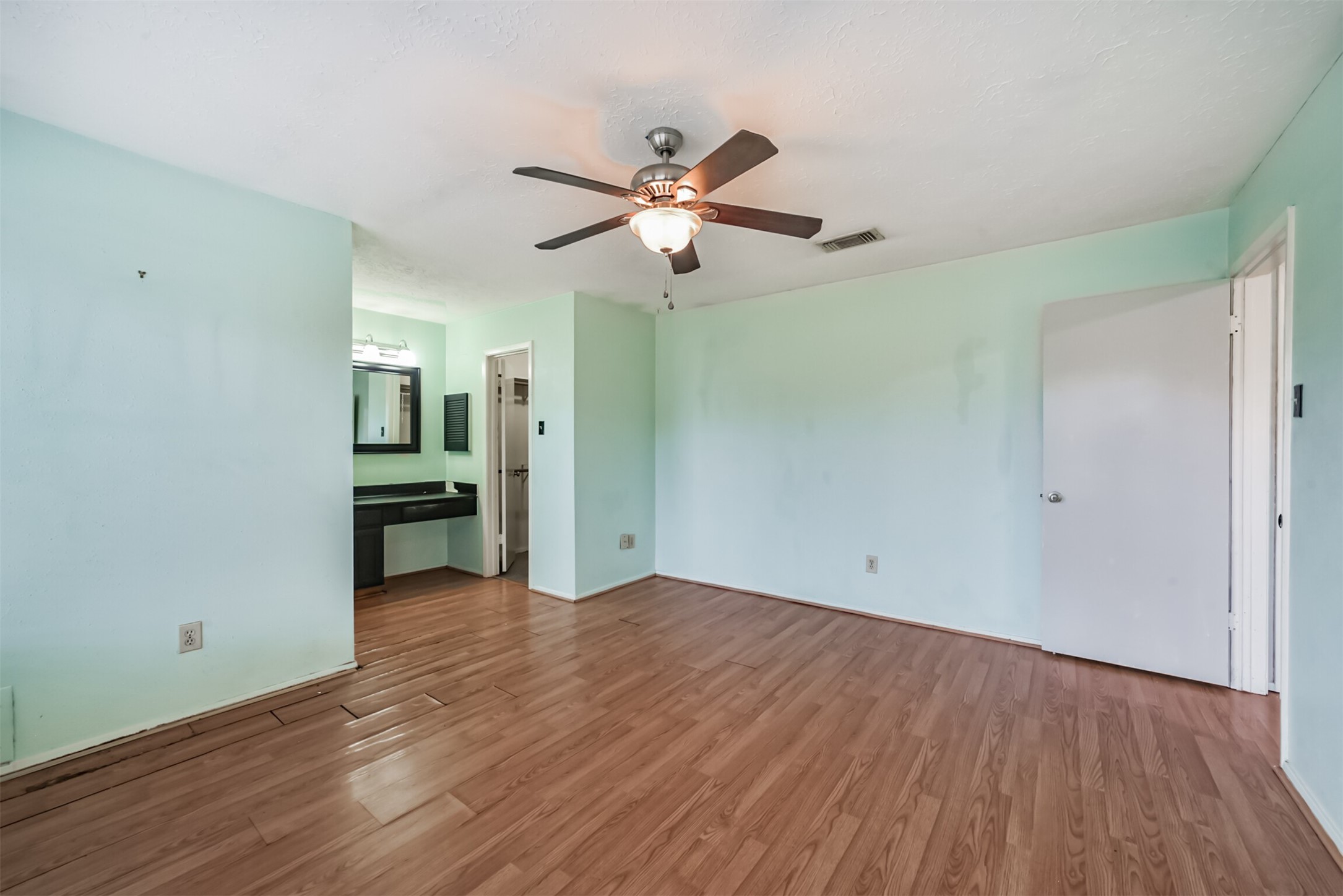 Spacious bedroom featuring laminate flooring, soft mint walls, a ceiling fan, and an attached vanity area for convenience.