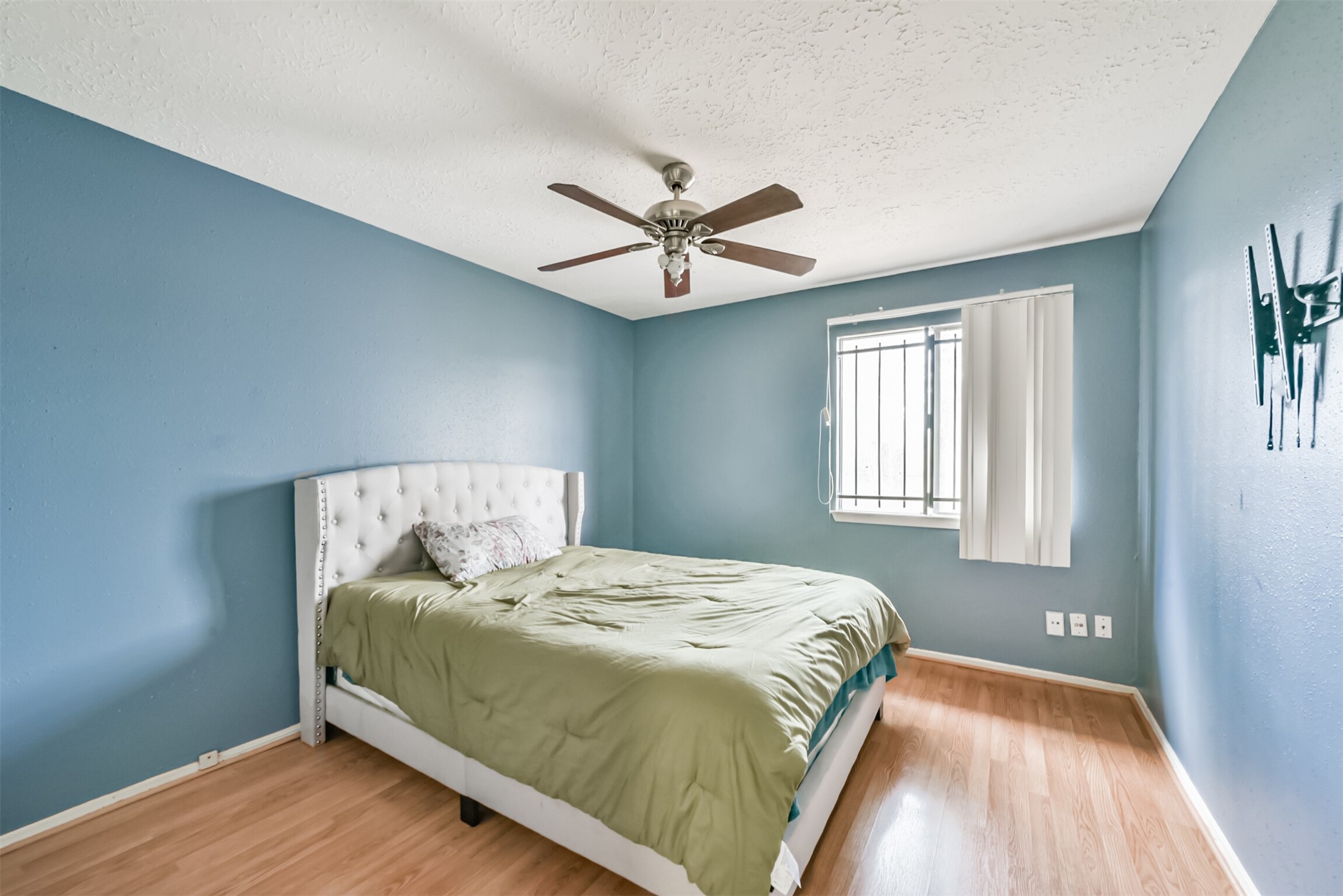 Charming bedroom with soothing blue walls, featuring a cozy bed and a ceiling fan for comfort. Natural light streams in through a window with vertical blinds, complementing the warm wood flooring.