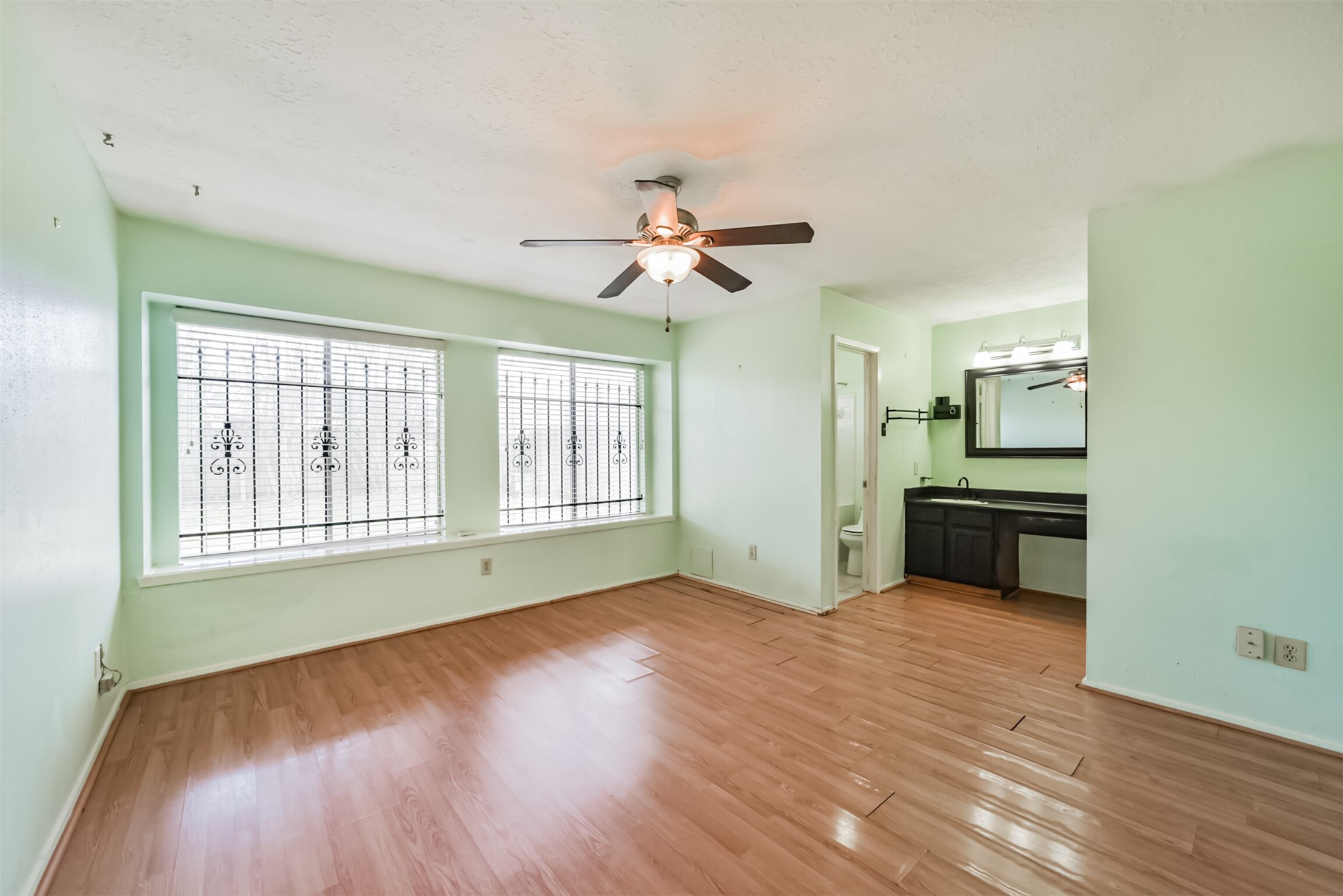 Spacious room with large windows featuring decorative ironwork, light mint green walls, and wood laminate flooring, complemented by a ceiling fan and a convenient vanity area.