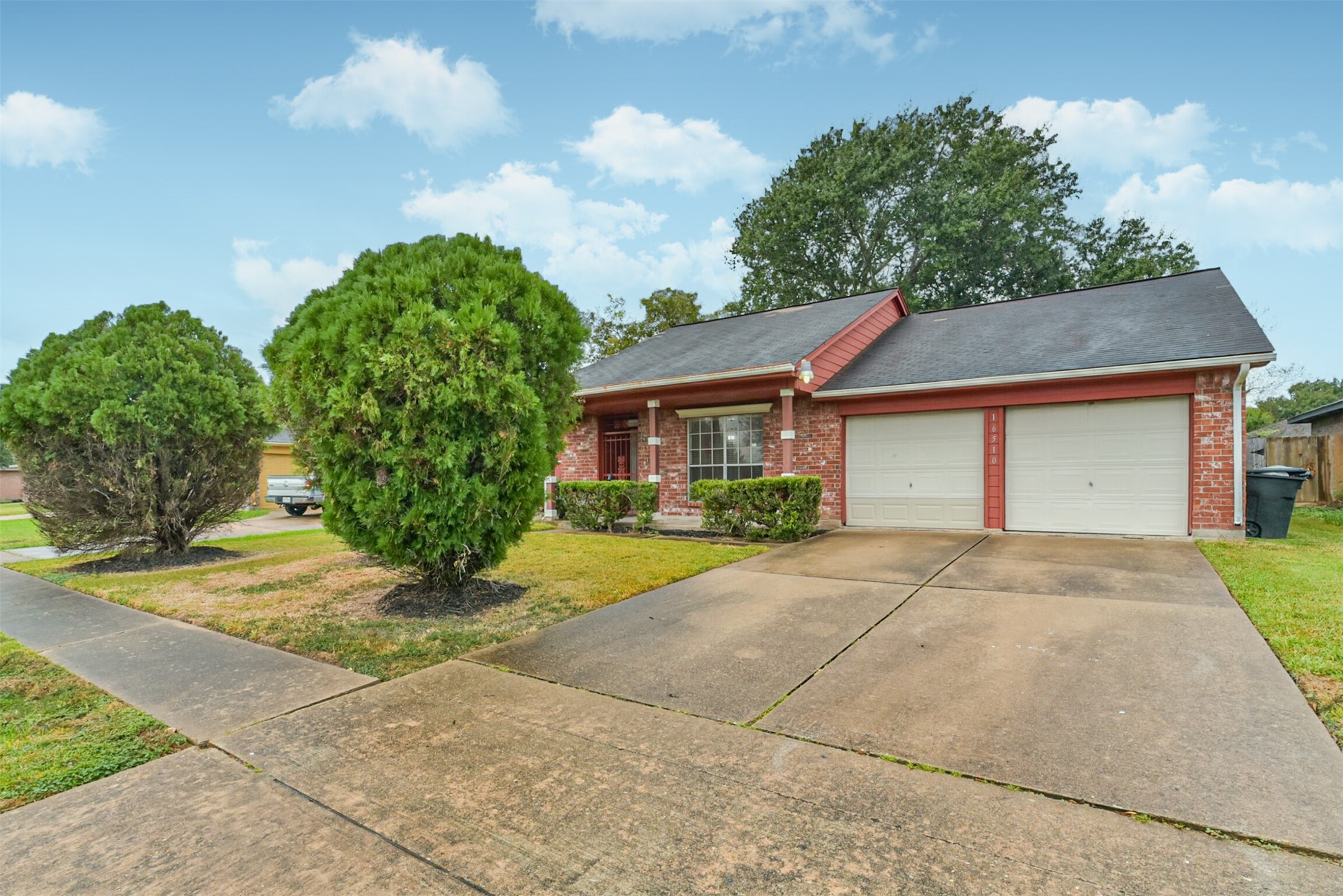 Charming brick home with a welcoming porch, dual garage doors, and a spacious driveway. Lush greenery and a well-maintained lawn enhance its curb appeal.