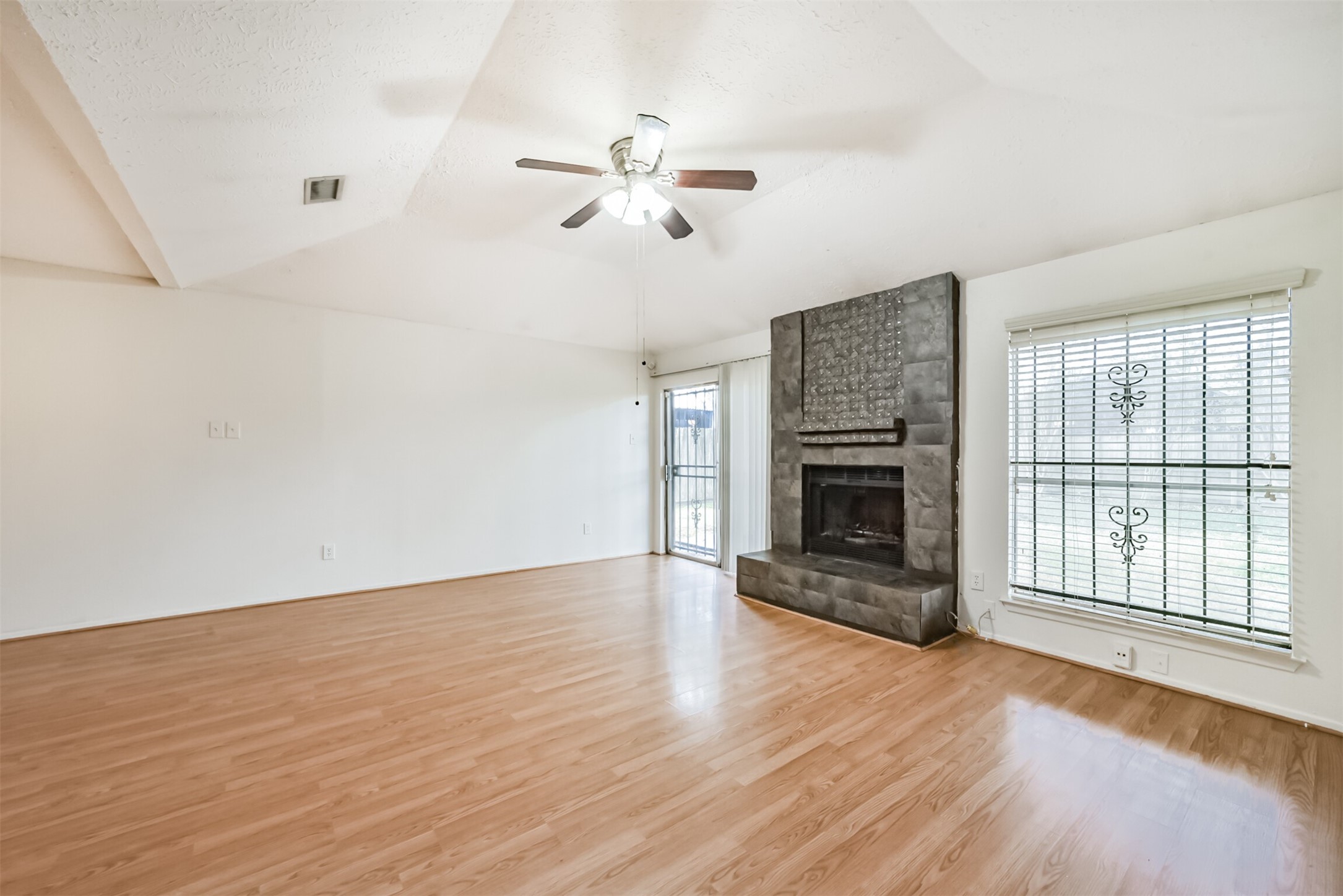 Spacious living room featuring elegant wood flooring, a modern ceiling fan, and a striking stone fireplace, accented by large windows for abundant natural light.