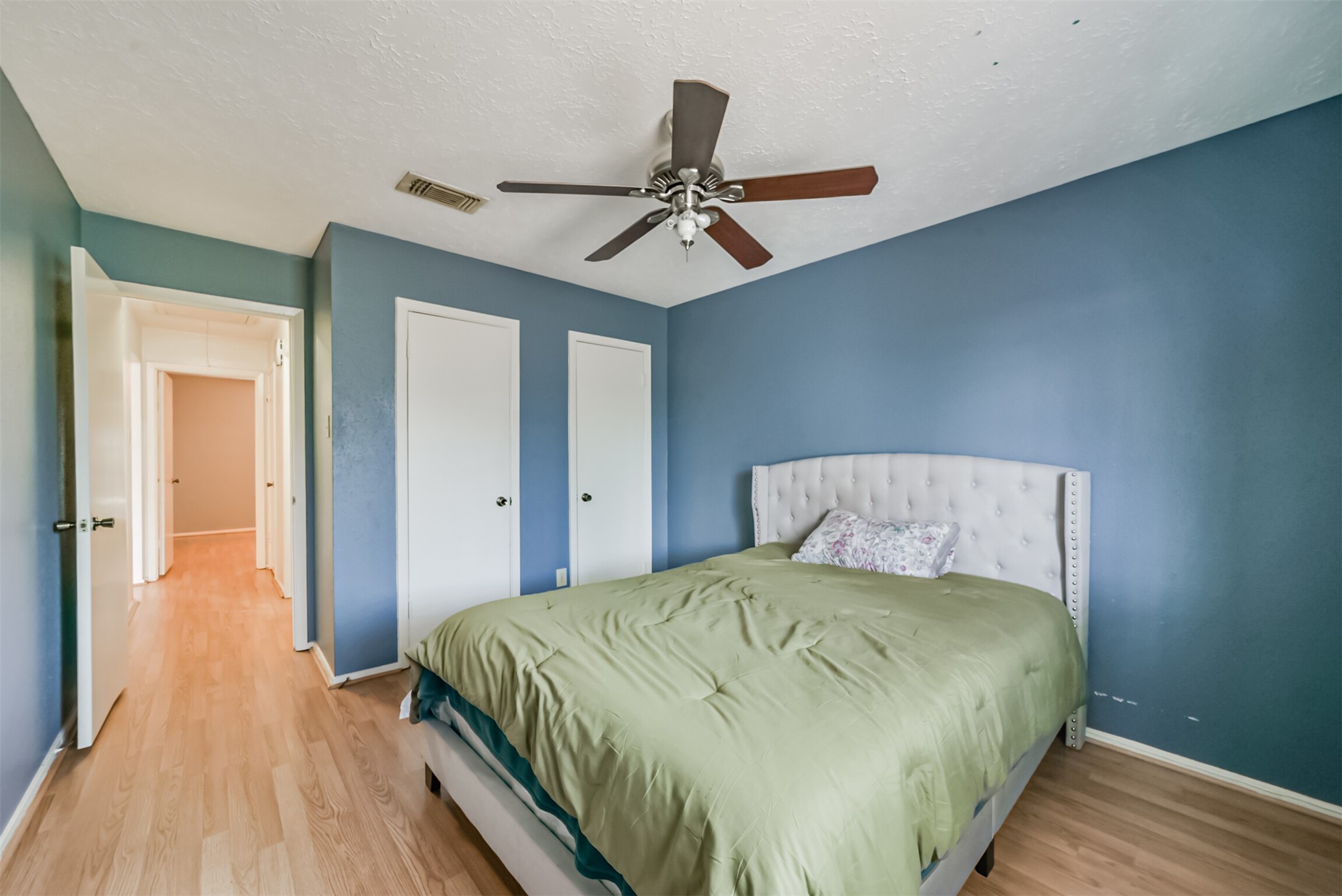 Cozy bedroom featuring a stylish tufted headboard, elegant ceiling fan, and warm wood flooring, accented by serene blue walls.