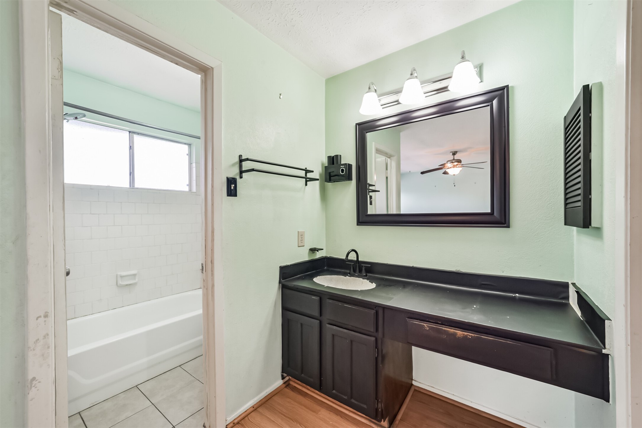 Charming bathroom with a modern black vanity, spacious mirror, and bright lighting, leading to a tiled bathtub area with natural light.