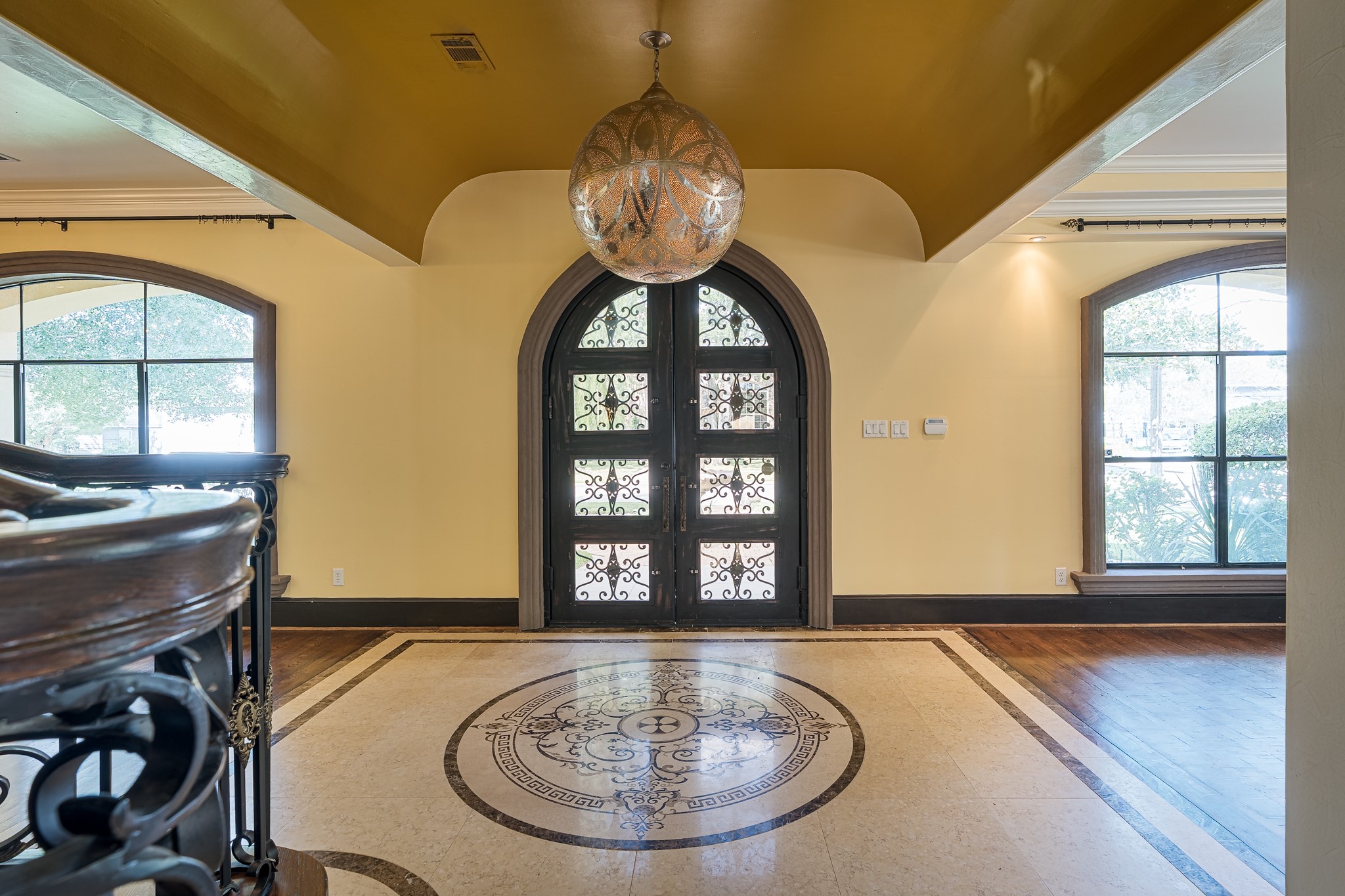 Elegant foyer with a stunning solid wrought-iron front door, ornate chandelier, and intricate tile floor design, complemented by large arched windows for abundant natural light.