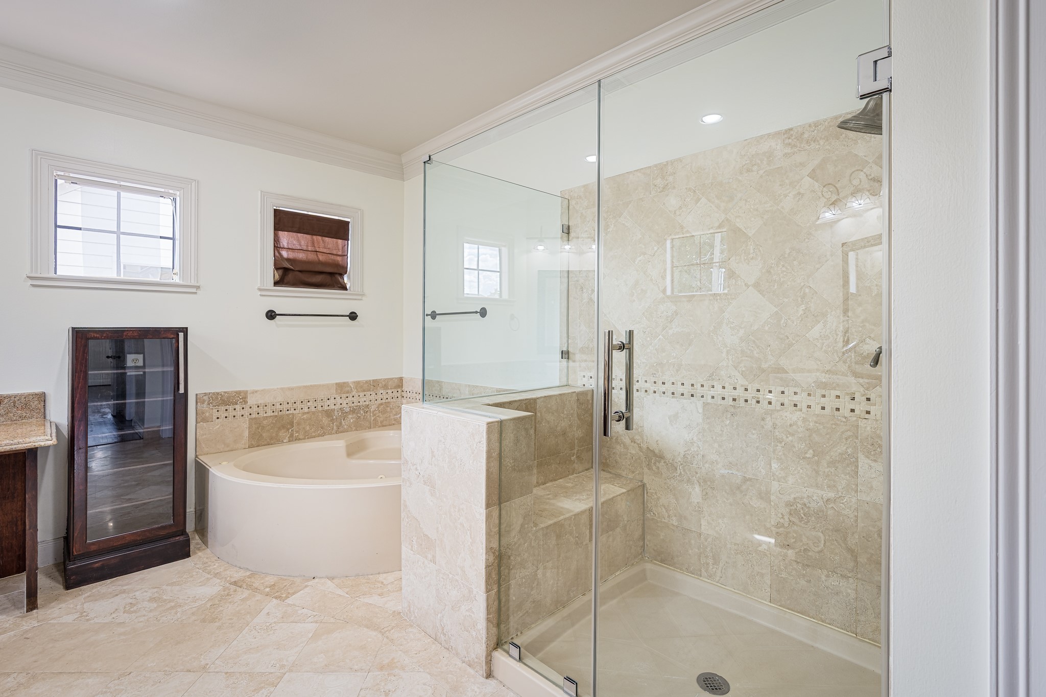 Elegant bathroom featuring a spacious glass-enclosed shower with travertine tile, a luxurious soaking tub, and ample natural light from the windows.