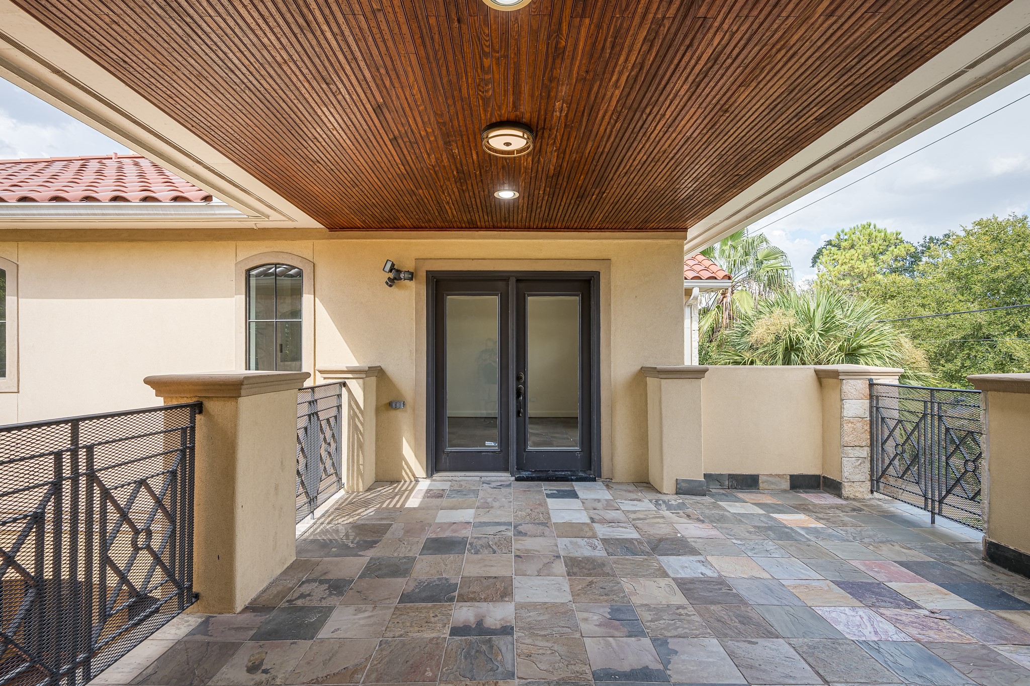 Spacious covered patio outside of the primary bedroom with elegant stone tile flooring, wood-paneled ceiling, and stylish iron railings, perfect for outdoor relaxation.