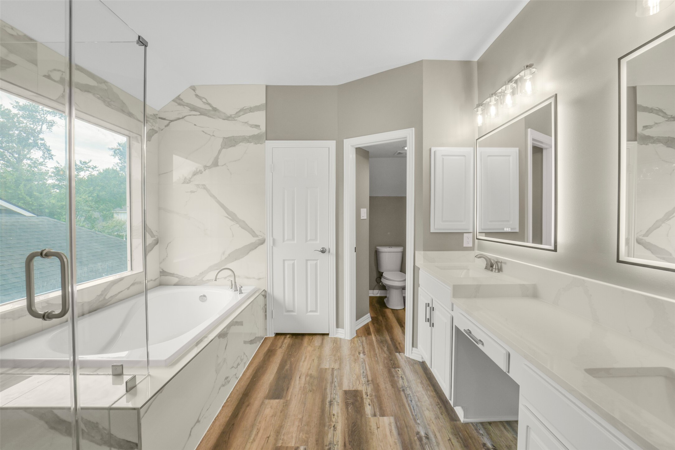 The spa-like primary bath features dual sinks with a vanity, a deep soaking tub, a glass enclosed walk-in shower, and a private commode area.
