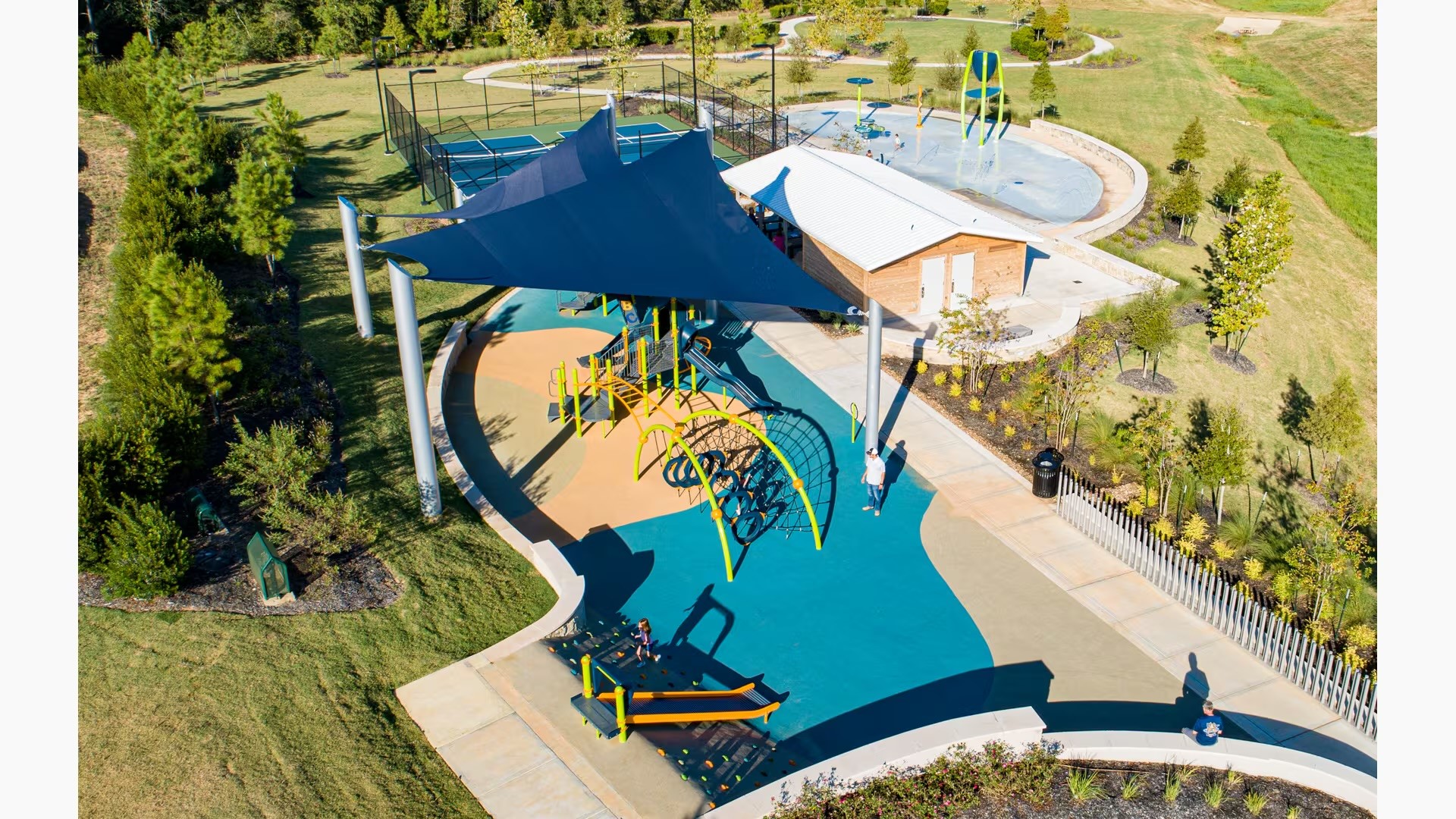 Experience vibrant outdoor living with this community park featuring a modern playground, shaded seating, tennis courts, and a splash pad, all surrounded by lush greenery.