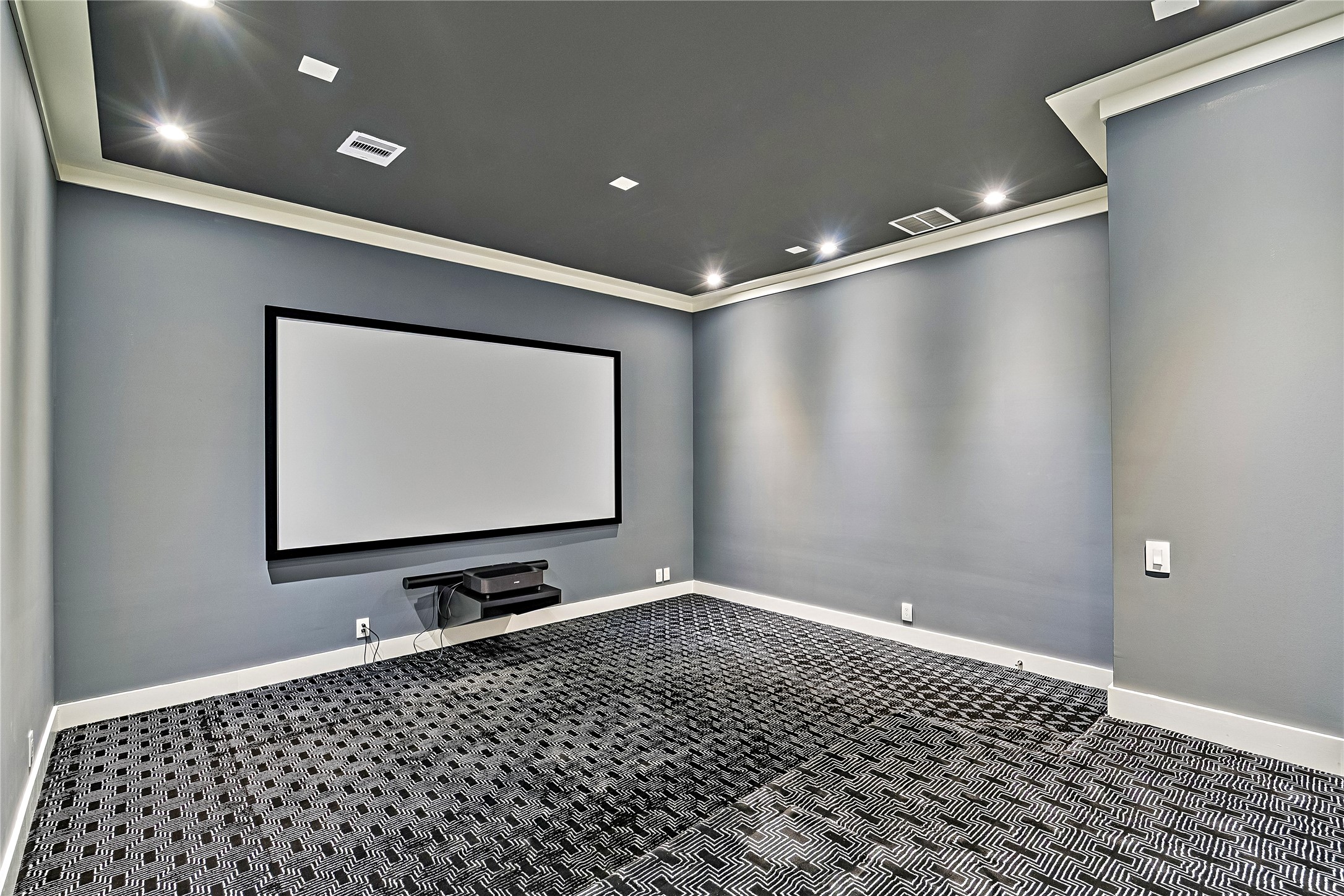 A separate media room featuring a large projection screen, dark ceiling treatment, recessed lighting, and patterned carpeting. Designed for private movie screenings and entertainment away from the home's principal living areas.